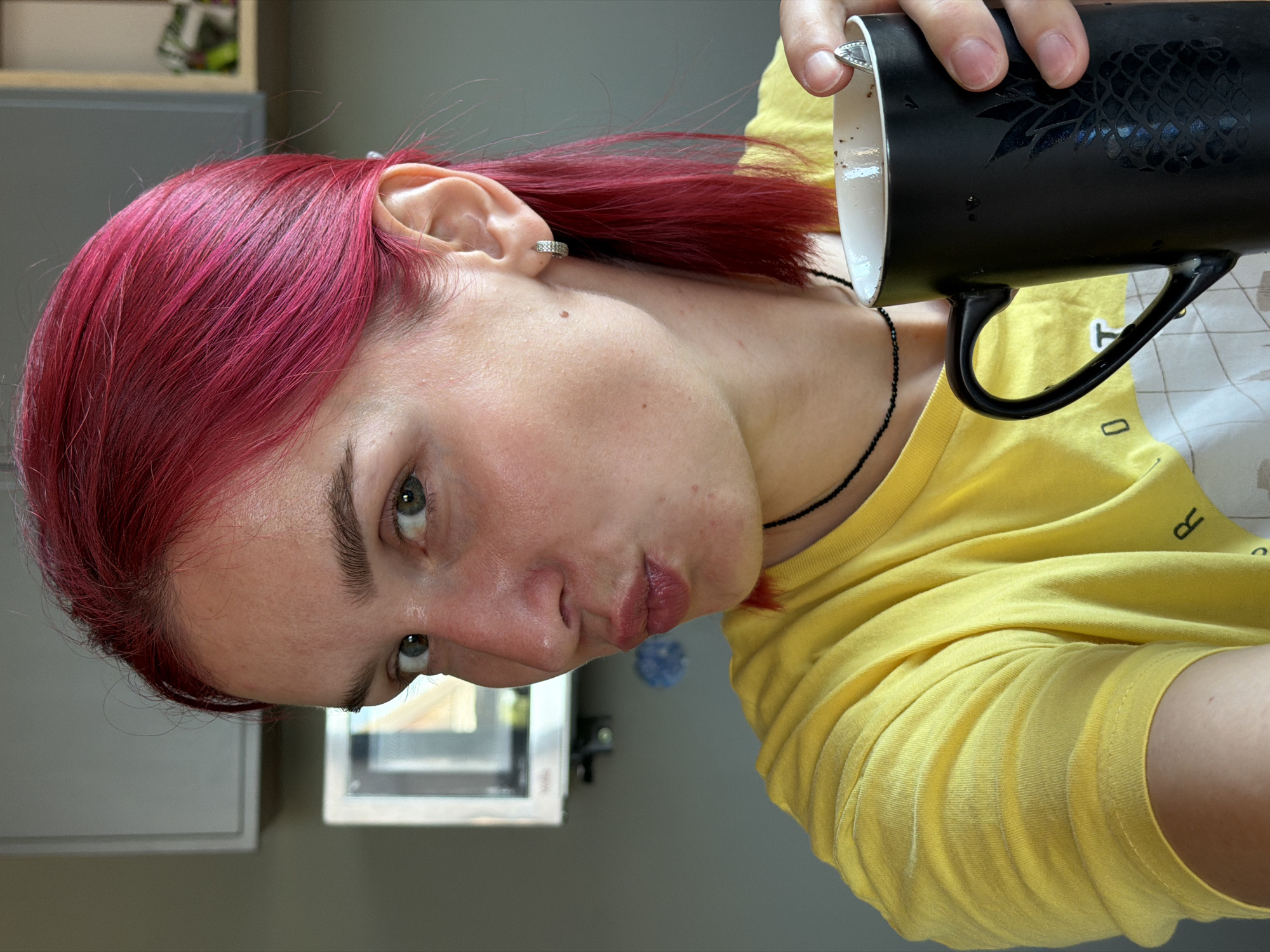 A close-up shot captures a young woman with vibrant pink hair indoors, holding a black mug with a pineapple design. She is wearing a yellow t-shirt and a thin black necklace. Her expression is a playful pout, with her lips pursed. She is looking directly at the camera with her left eye visible. In the background, a microwave is mounted on a grey wall. The overall impression is a casual, personal moment. The specific location in Chaikovsky, Russia, cannot be determined from the visual information provided in the image.