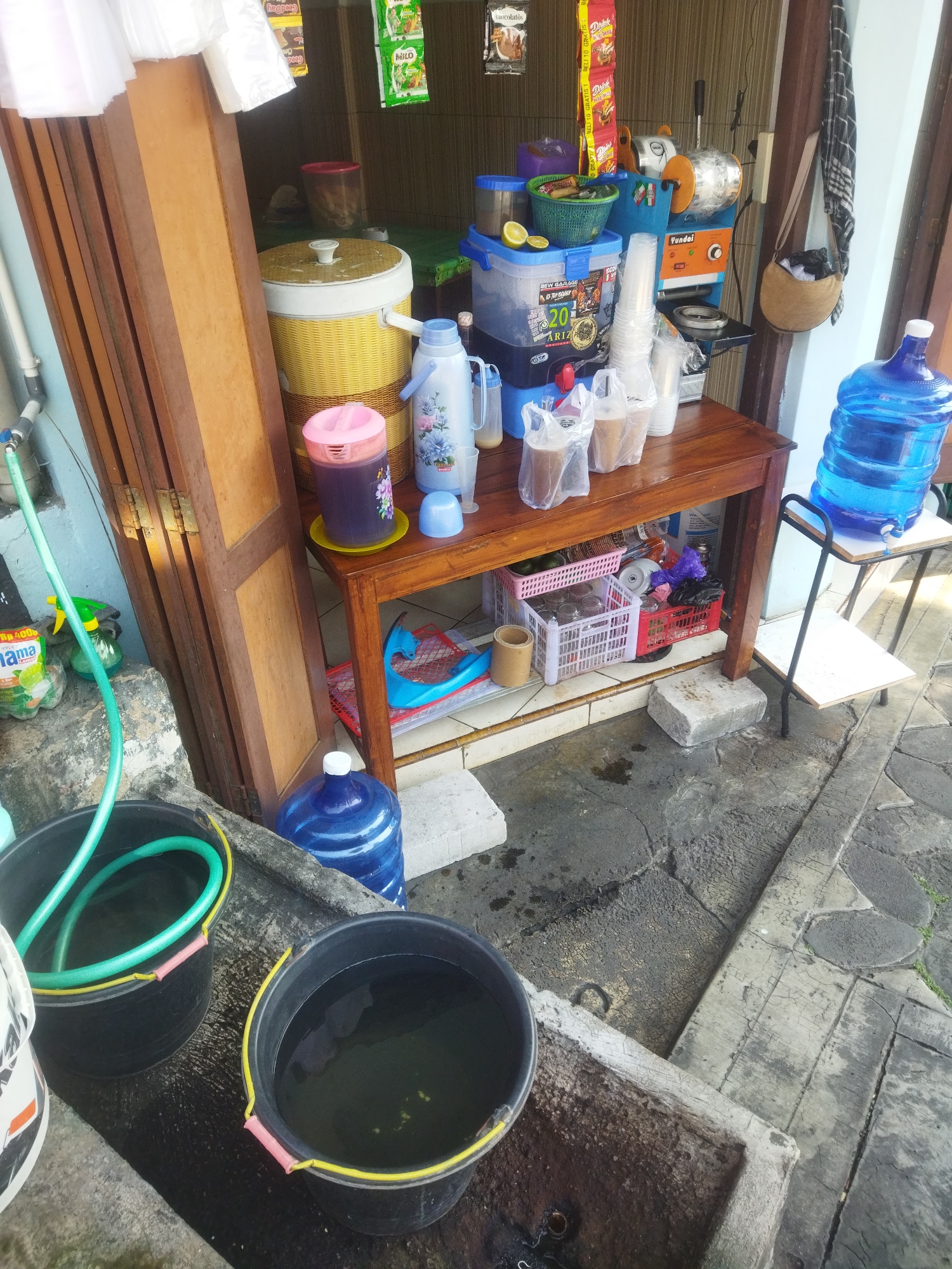 This is a medium shot, eye-level photograph of a street-side beverage stand in Wonogiri, Indonesia. The stand appears to be a small, makeshift operation located near a building. The primary focus is on a wooden counter laden with various items for preparing and serving drinks.
To the left of the counter, there is a large, yellow, woven cooler or ice chest with a white lid. Next to it is a tall, purple beverage dispenser with a floral pattern and a pink lid. Further to the left, a green hose is coiled inside a black bucket, with another black bucket beside it also containing water. A large blue water jug with a white cap is visible to the right of the counter.
On the counter itself, there are several items including a blue liquid dispenser with a dispenser tap, clear plastic cups, and plastic bags filled with beverages. Behind these, a silver and orange ice-shaving machine is mounted. Various packets of powdered drinks and snacks are displayed hanging above the counter. Two lemons are visible in a blue basket on top of the liquid dispenser. A dark-colored woven bag with a strap is hanging on the right side of the frame.
The ground in front of the stand is paved with rough concrete and shows signs of wear and occasional puddles. The background shows the side of a building with a wooden door. The overall impression is of a functional, if somewhat cluttered, beverage stall operating in an urban outdoor environment. There are no people visible in the image.