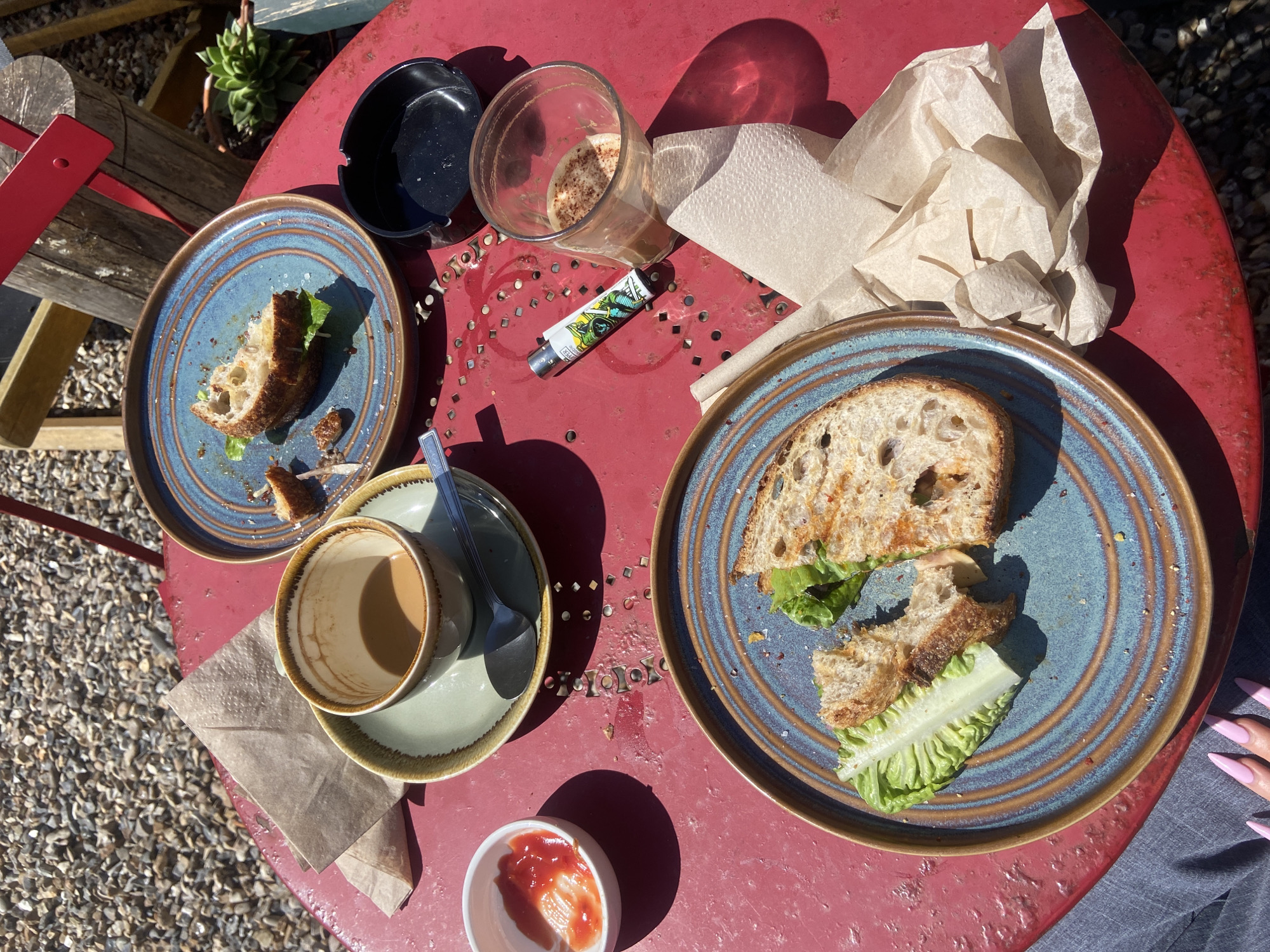 The image depicts a partially consumed meal on a red, round metal table. The setting appears to be outdoors, with a gravel surface visible in the background.
The meal consists of two plates of sandwiches, with one plate having only remnants of a sandwich left. Both plates are blue with brown rims. Next to one plate sits a matching cup and saucer with a spoon. The cup is mostly empty. There is a lighter, an empty glass, and crumpled paper napkins on the table, suggesting the meal is finished. A small dish containing a red sauce is also present. Part of a chair can be seen to the left. The shadows indicate sunny conditions. It is not possible to determine the location is in London, United Kingdom, or determine if any specific people are present.