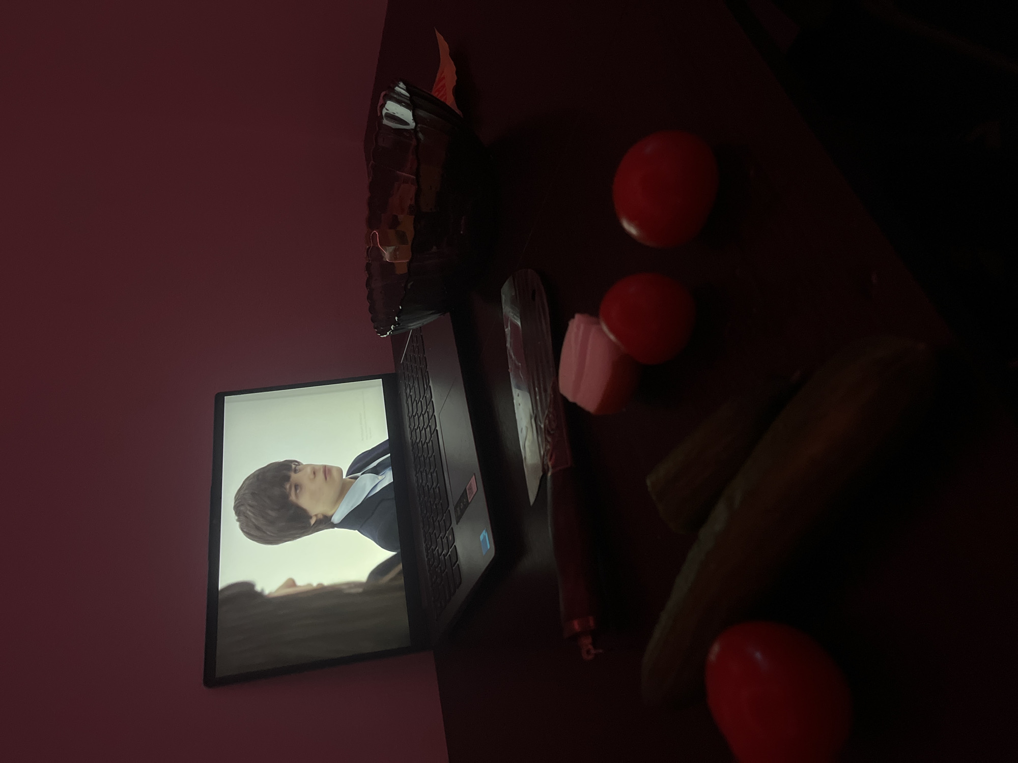 A dimly lit scene presents a laptop displaying a person on its screen, situated on a table amidst a collection of items. The laptop shows a male figure with dark hair and a blue shirt, possibly in a scene from a video or movie. Adjacent to the laptop are two whole tomatoes, a pink object, and what appears to be a knife. There are also two cucumbers and another tomato towards the right side of the table. To the left, there is a vase on the table. Given the objects' positions and the presence of what appears to be food items and utensils, the scene might represent a casual setup with a meal preparation and entertainment. The image is taken with the camera oriented in a top-down view. The photo was not taken in Saint Petersburg, Russia.