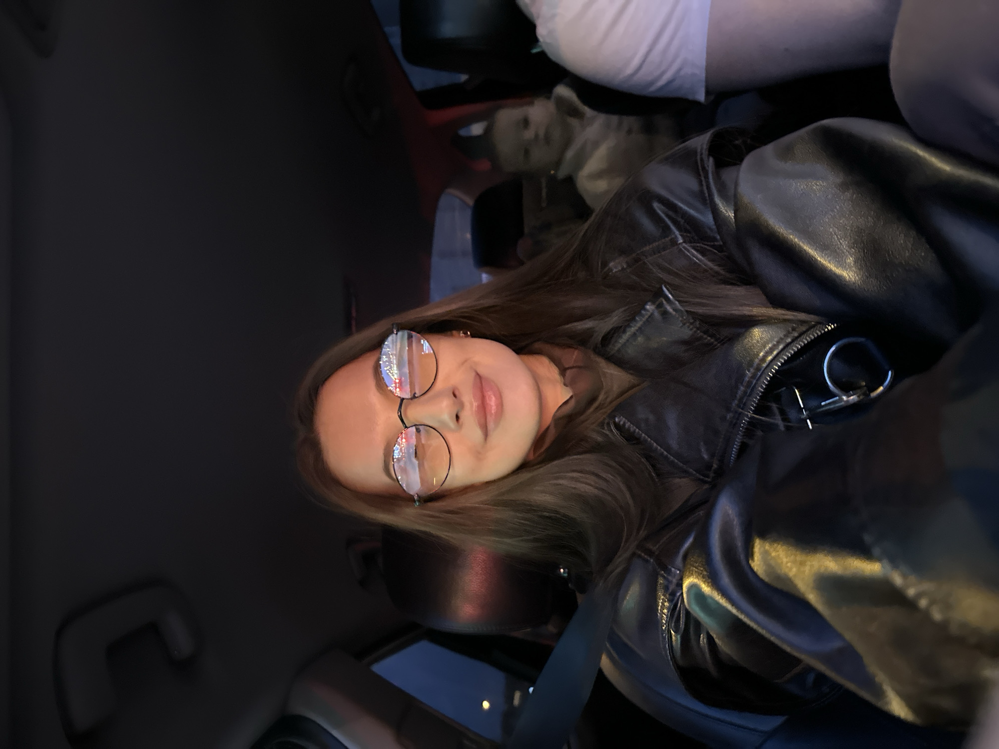 A person wearing round glasses and a black leather jacket is sitting in a car. The person is facing the camera with a slight smile. The car appears to be in motion at night, as reflected in the glasses. Another person is in the backseat. The location is Yekaterinburg, Russia, based on the reflection in the glasses showing a street scene with lights.