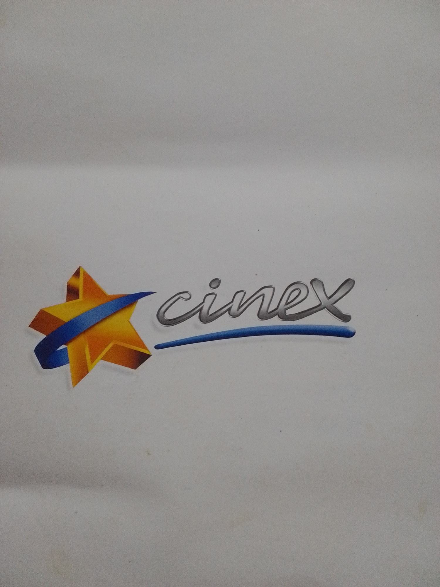 This image shows the logo for "cinex" a Venezuelan cinema chain. The logo features a stylized orange star with a blue ribbon wrapped around it, positioned to the left of the word "cinex" which is written in a silver, cursive font. The background is a plain, light gray surface. The logo is centered horizontally in the frame. There is no indication of the city of La Tagua, Venezuela within the image itself.
