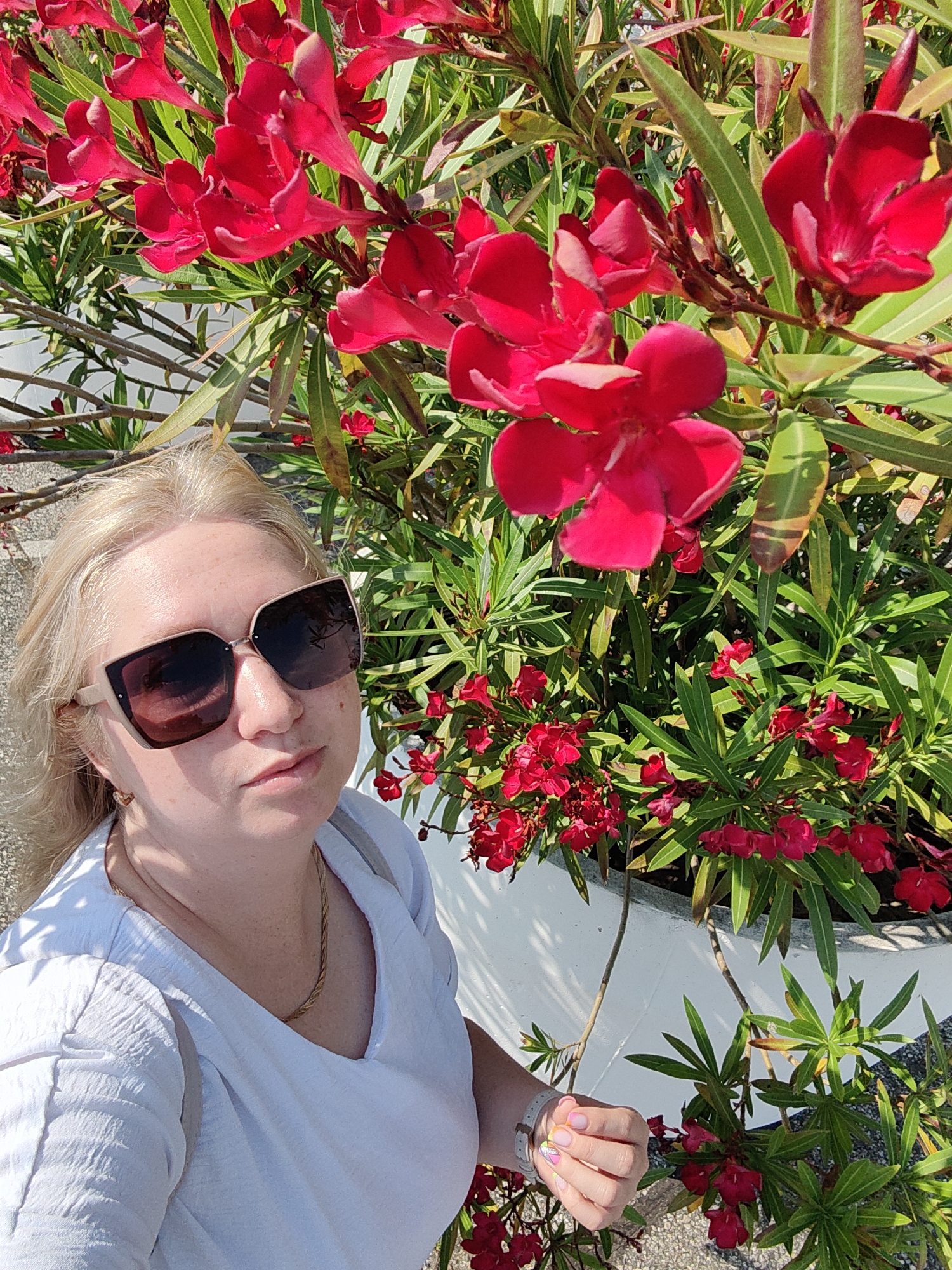 A blonde woman with fair skin and freckles, wearing oversized sunglasses and a white v-neck shirt, takes a selfie in front of a bush with vibrant red flowers and green leaves. Her left hand is extended forward, showing painted nails. She is wearing a gold chain necklace. The background appears to be outdoors with a white planter and a light-colored, textured surface beneath her feet. The lighting suggests a sunny day. The location is identified as Sirius, Russia.