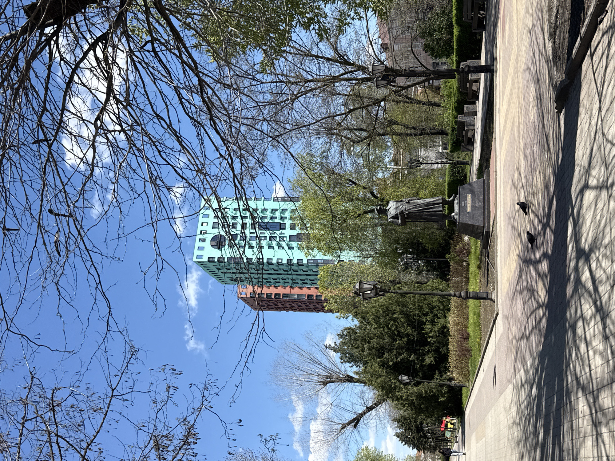 A daytime shot of Rostov-on-Don features a statue in a park, partially obstructed by a streetlamp and trees, with a modern building visible in the background. The structure has a distinct two-tone color scheme, with the upper portion being green and the lower portion being brick-colored. The sky is blue with some scattered white clouds. Bare tree branches dominate the left side of the frame, creating a visual contrast with the building. Pigeons are on the paved pathway beside the statue.