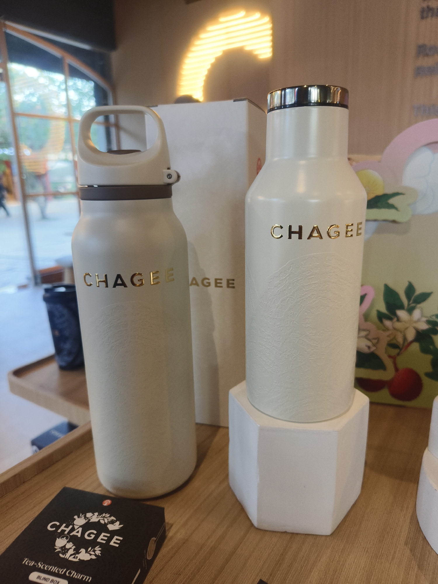Two cream-colored CHAGEE brand water bottles are displayed on a wooden surface. The bottle on the left has a brown band around its neck and a cream-colored handle. The bottle on the right sits on a white hexagonal stand and has a gold-colored rim. Both bottles have the CHAGEE logo printed in gold. In the foreground, a black box with white text and illustrations reads "CHAGEE Tea-Scented Charm BLIND BOX." In the background, out-of-focus elements include a window with greenery and a large yellow light fixture. The overall setting appears to be a retail environment.