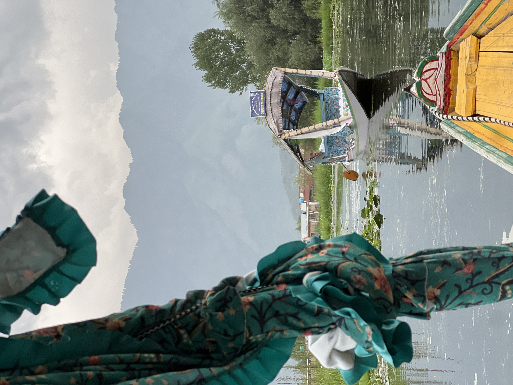 This is a medium shot, taken from a boat on Dal Lake in Srinagar, India, looking across the water towards another boat and the distant mountains. In the foreground, a large piece of teal fabric with a floral pattern is draped, obscuring the left side of the image. This fabric appears to be part of a canopy or covering.
In the mid-ground, a white shikara, a traditional Kashmiri wooden boat, is being rowed by a man wearing a white shirt. The shikara has a canopy with decorative blue and white patterned fabric lining its interior and a sign that reads "NEW ABDULLAH PALACE DELUXE SHIKARA". The boat is surrounded by lily pads and other aquatic vegetation.
In the background, a line of wooden houses on stilts is visible along the shore, with trees and lush green vegetation. Beyond the houses, a range of hazy, blue-tinted mountains forms the horizon, under a cloudy sky. The water reflects the sky and the surrounding elements, creating a serene atmosphere. Another boat, predominantly yellow with red and green accents, is partially visible in the lower right foreground, suggesting the photographer is also on a boat.