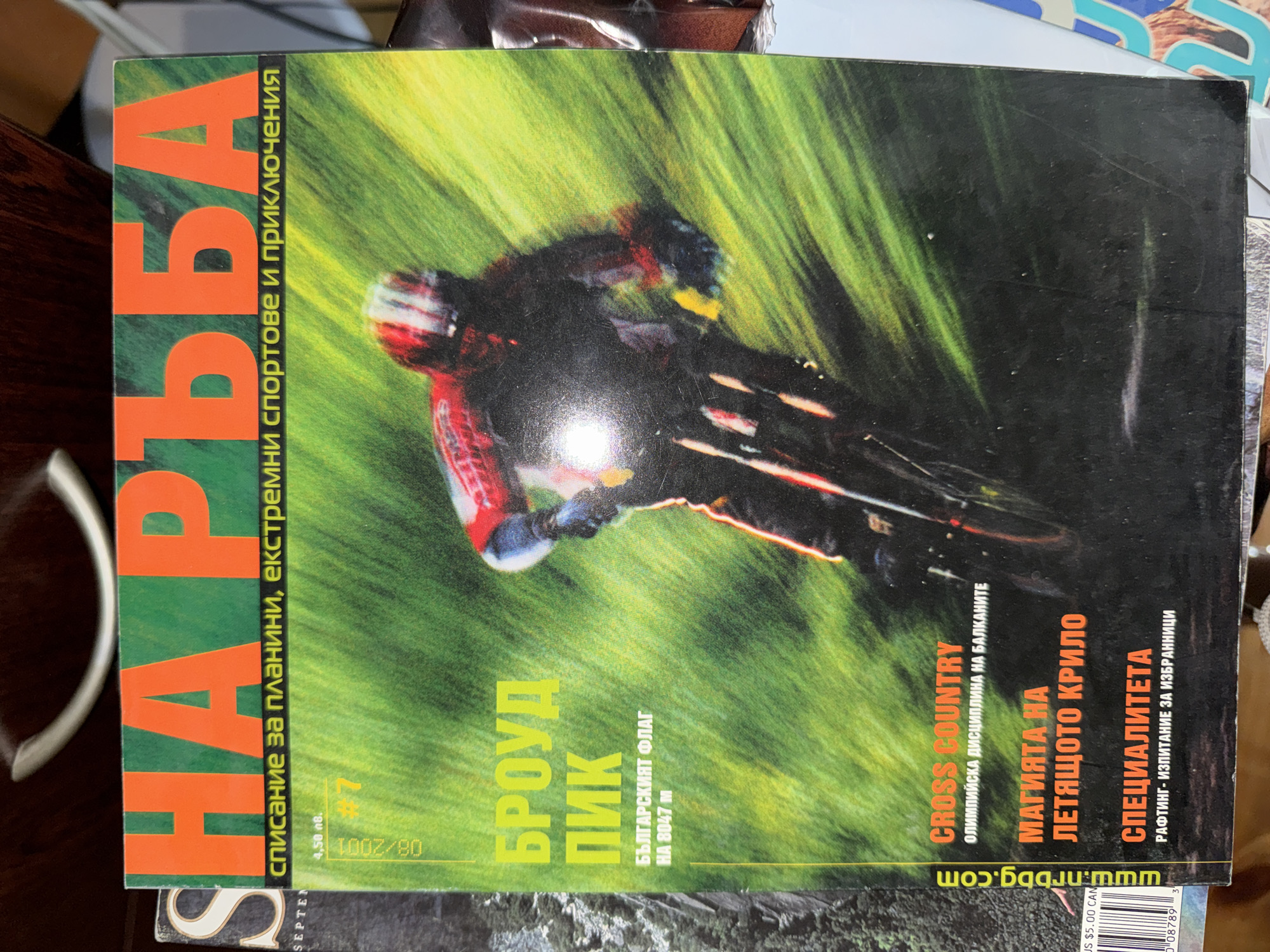 The image displays the cover of a Bulgarian extreme sports and adventure magazine titled "НА РЪБА" (On the Edge). The main subject on the cover is a mountain biker in action, captured in mid-turn or descent. The biker wears a red and white helmet and cycling gear, with vibrant colors that pop against the background. The mountain bike is dark, with visible details suggesting it's designed for rugged terrain. The background is a dynamic, blurred green, indicating high speed or motion through grass or foliage, creating a sense of exhilaration and movement.
The magazine, issue #7 from August 2001, caters to enthusiasts of mountains, extreme sports, and adventures, as stated in its slogan "списание за планини, екстремни спортове и приключения." Visible text highlights several articles:
- "БРОУД ПИК" (Broad Peak), featuring "БЪЛГАРСКИЯТ ФЛАГ НА 8047 м" (The Bulgarian Flag at 8047 m), suggesting a Bulgarian expedition.
- "CROSS COUNTRY," described as an "ОЛИМПИЙСКА ДИСЦИПЛИНА НА БАЛКАНИТЕ" (Olympic Discipline in the Balkans).
- "МАГИЯТА НА ЛЕТЯЩОТО КРИЛО" (The Magic of the Flying Wing), likely about paragliding or hang gliding.
- "СПЕЦИАЛИТЕТА" (Specialty) on "РАФТИНГ - ИЗПИТАНИЕ ЗА ИЗБРАННИЦИ" (Rafting - A Test for the Chosen Ones).
The cover also shows pricing information: 4.50 BGN and $5.00 CAN, and the website "www.nrbbg.com". The overall impression is one of high-energy, outdoor adventure and exploration, likely targeting a readership interested in thrill-seeking activities within Bulgaria and internationally, given the general context of Sofia, Bulgaria as a hub for such publications.