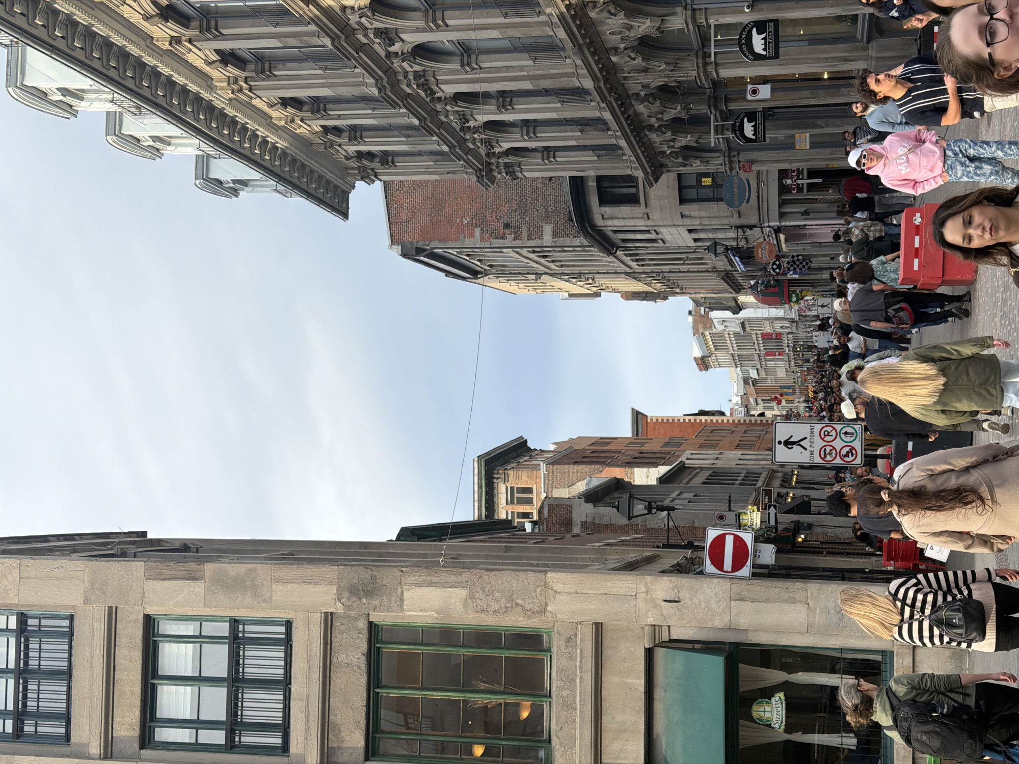 A street scene in Montreal, Canada, is captured, densely populated with pedestrians and framed by building exteriors on either side. The architectural styles vary, with some buildings exhibiting ornate detailing. Street signs and business names, such as "Suzette" and "Galerie d'Art Inuit," are visible. Numerous individuals are seen strolling, some wearing casual attire like a pink hooded sweatshirt and patterned pants. A woman is pulling a red suitcase on wheels. The sky is partially visible between the buildings. A pedestrian zone sign is visible that indicates, in French, that pedestrians must use this route. A red stop sign is seen, too.