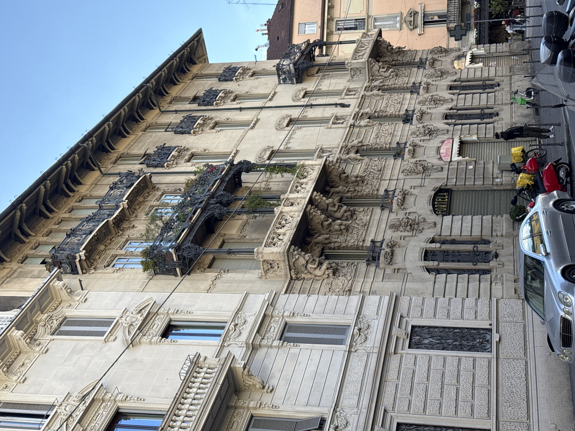 The image depicts the facade of Palazzo Castiglioni, a historical building located in Milan, Italy. The building is characterized by its ornate Art Nouveau style, evident in the sculpted details, wrought-iron balconies, and decorative window frames. The facade is predominantly light-colored stone. Some balconies have plants.
In the foreground, a sidewalk features a parked silver car and several parked motorbikes, some with food delivery boxes. A person is walking on the sidewalk near the motorbikes. Other buildings are visible in the background. The sky is clear and blue.