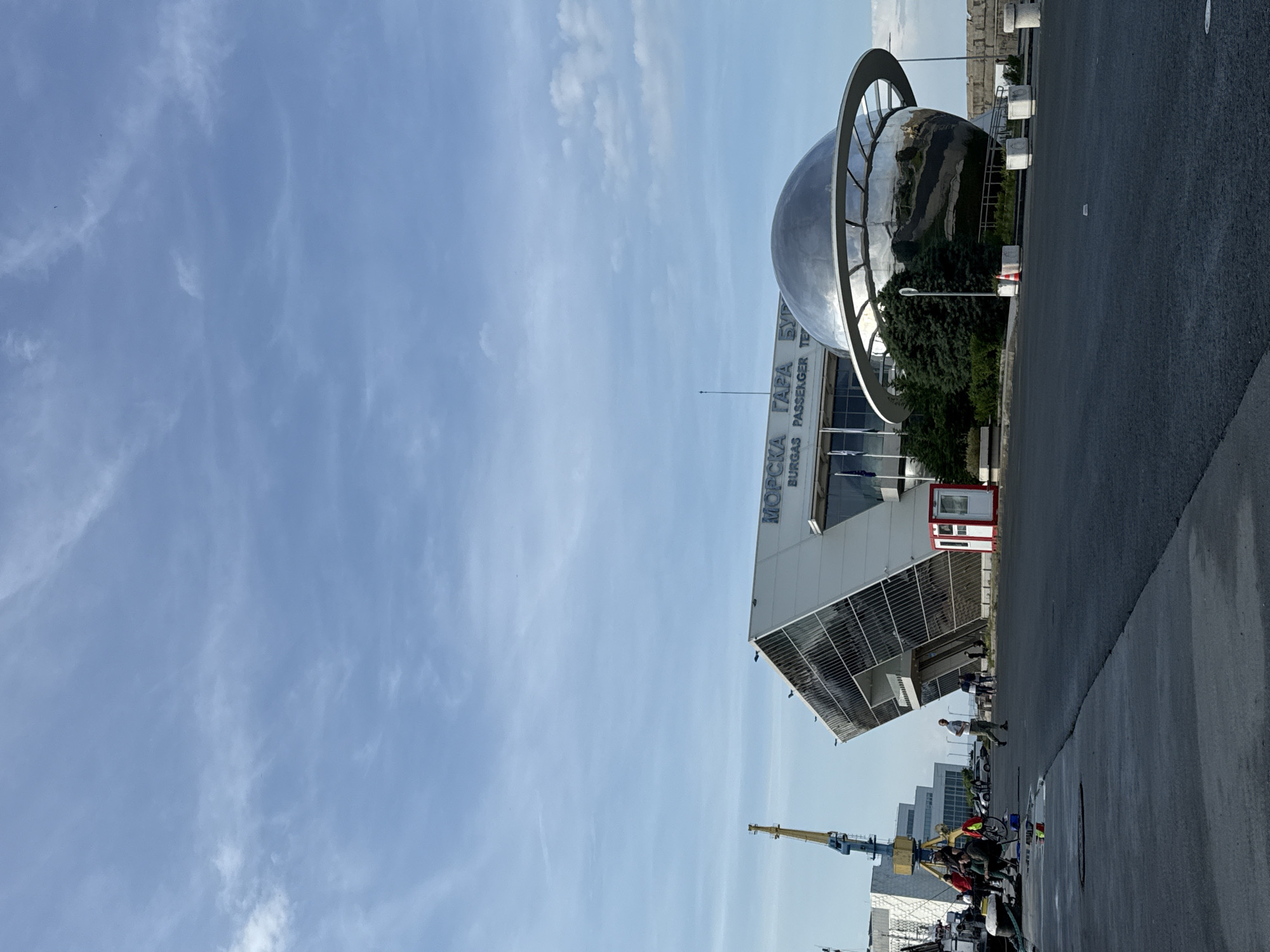 The image shows the Burgas Passenger Terminal in Burgas, Bulgaria. The building is white with a modern design and features the inscription "МОРСКА ГАРА БУРГАС PASSENGER TERMINAL". A large, silver, spherical sculpture surrounded by a ring is located in front of the terminal. The sky is blue with wispy clouds. To the right, there is a paved area that appears to be a street or parking area. In the distance, there are industrial cranes and other buildings. People are visible on the paved area near the terminal.