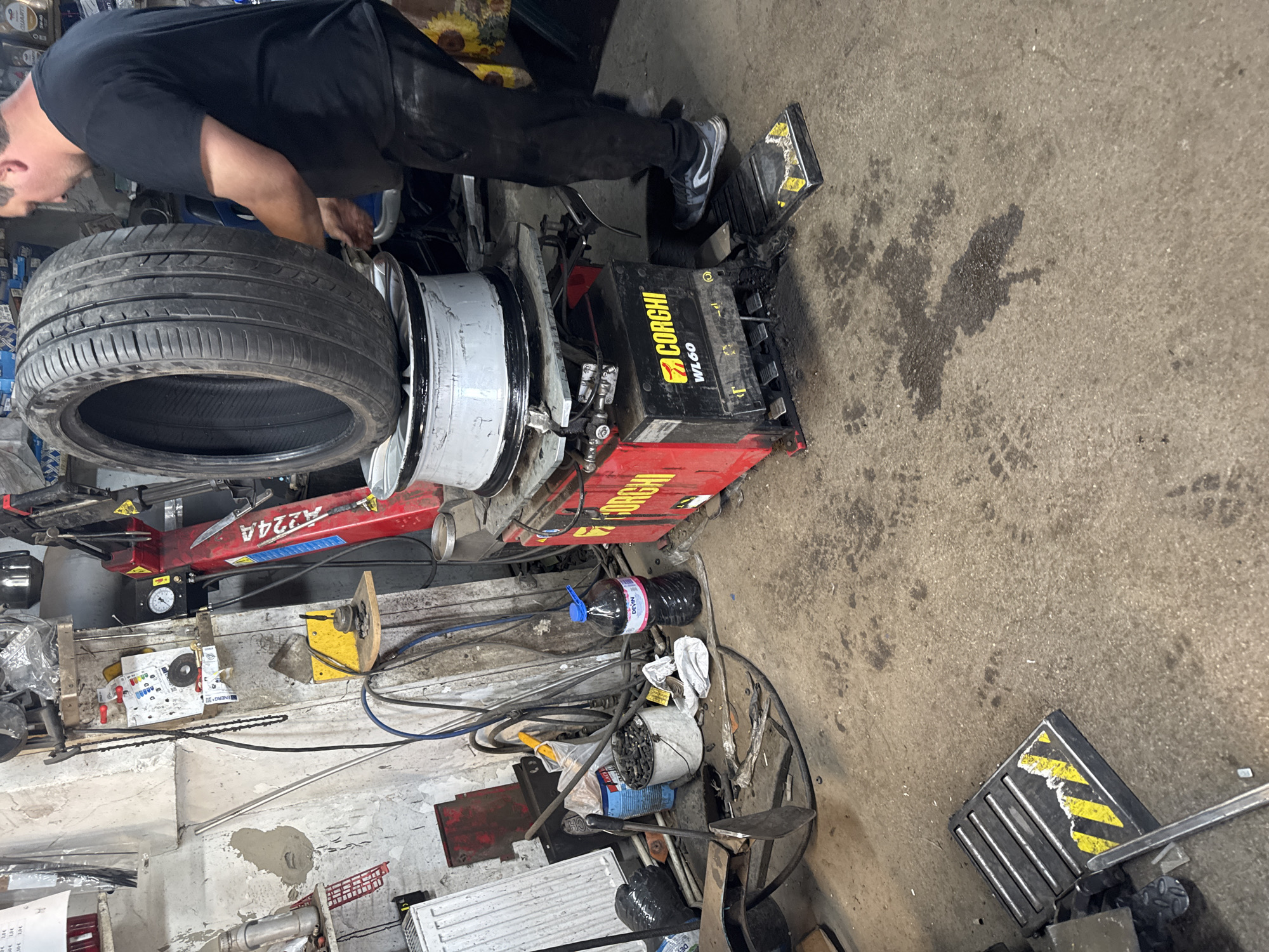 In a garage in Batanovtsi, Bulgaria, a man is working on a tire. The tire is mounted on a tire changing machine. The garage floor is concrete and stained. There are tools and equipment scattered around the work area. The tire changing machine is red and black and has the brand name "CORGHI". A red A224A is visible on the tire changing machine. The man is wearing a black shirt and black pants. He is standing next to the machine and appears to be using a tool to work on the tire.