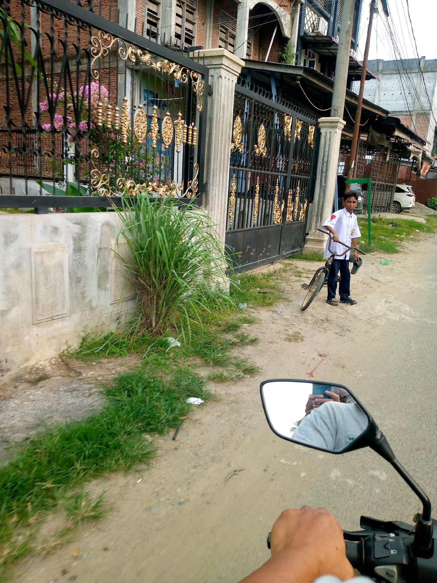 The image depicts a street scene in Keudegeudong, Indonesia. A young boy, dressed in a white school uniform with a red emblem on the left sleeve and dark pants, stands on the side of a paved road holding a bicycle by its handlebars. He is also holding a pair of dark sandals in his left hand. The bicycle is dark in color with a red accent on the frame. Behind the boy is a black metal gate with ornate gold embellishments, which appears to be the entrance to a property. Beyond the gate, a multi-story building with brick and concrete construction is visible. To the left of the gate, a concrete wall with two rectangular indentations runs along the street, partially covered by lush green grass and a large clump of tall green reeds. Further down the road, a white car is parked. The perspective of the image is from someone riding a motorcycle, with a portion of the motorcycle's handlebar and a rearview mirror in the foreground. The mirror reflects the rider's arm and shoulder, wearing a light-colored t-shirt, and a blue mobile phone. The environment suggests an urban or semi-urban setting with buildings, a road, and vegetation.