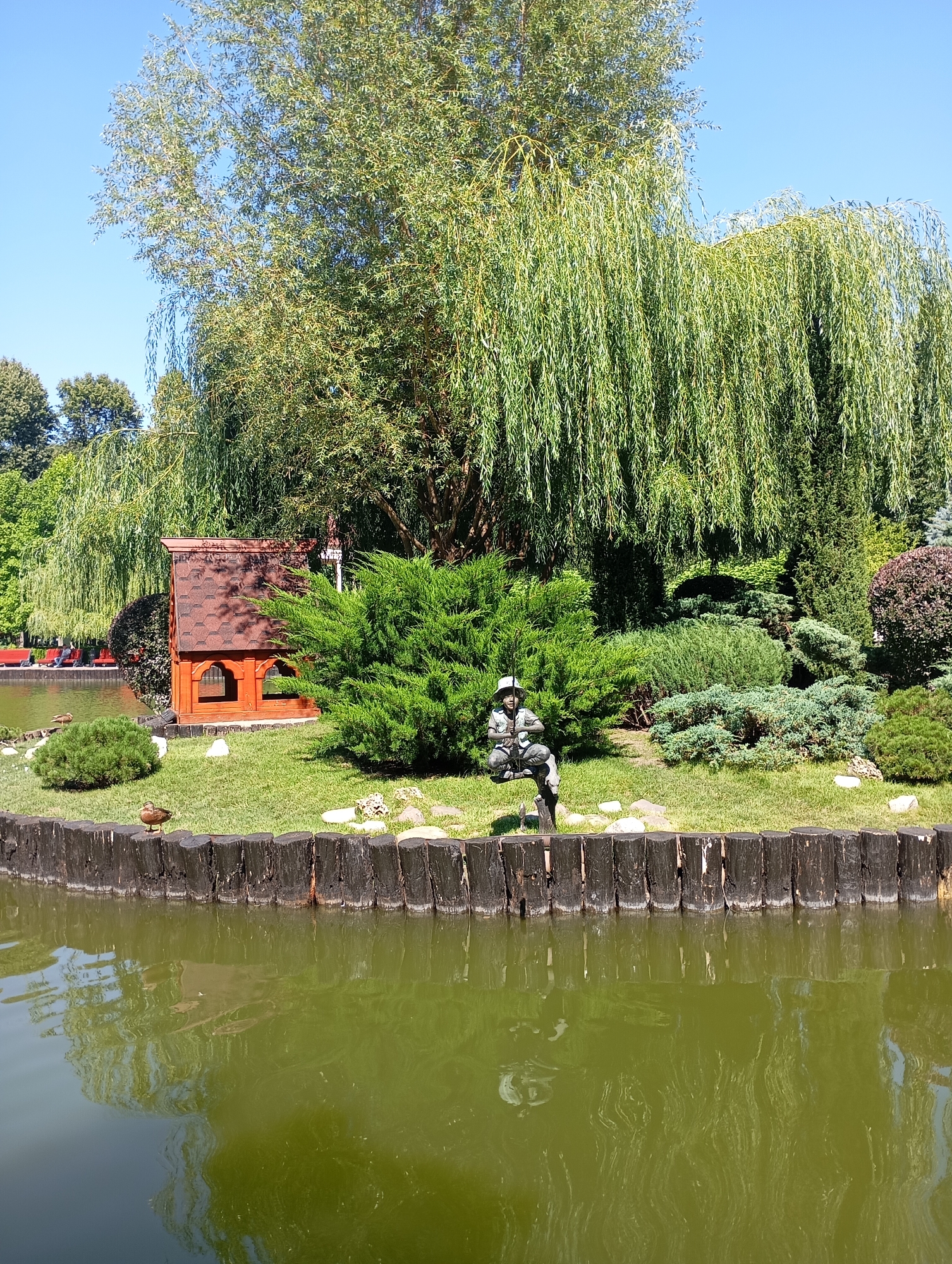 This is an outdoor, eye-level, medium shot of a picturesque park scene with a body of water in the foreground. The water is a greenish hue and reflects the surrounding trees and sky. A dark wooden fence made of vertical logs borders the water. On the grassy bank behind the fence, a small, rustic wooden structure with a shingled roof is visible, resembling a miniature house or shelter. Beside it, a dense green bush stands prominently. To the left of the bush, a small, round ornamental bush is planted. Further back, a statue of a person, possibly a fisherman wearing a hat, is seated on a low, rough-hewn perch. The statue is surrounded by various evergreen bushes and trees. A large weeping willow tree dominates the background, its long, drooping branches creating a curtain of green. A tall deciduous tree with a full canopy stands to the left of the willow. The sky is a clear, bright blue. A single duck is visible on the grass near the fence. In the distance, across the water, there are glimpses of red benches and more trees, suggesting this is part of a larger park. The overall impression is one of a tranquil, well-maintained garden or park area. This scene is located in the Kharkiv Zoo in Kharkiv, Ukraine.