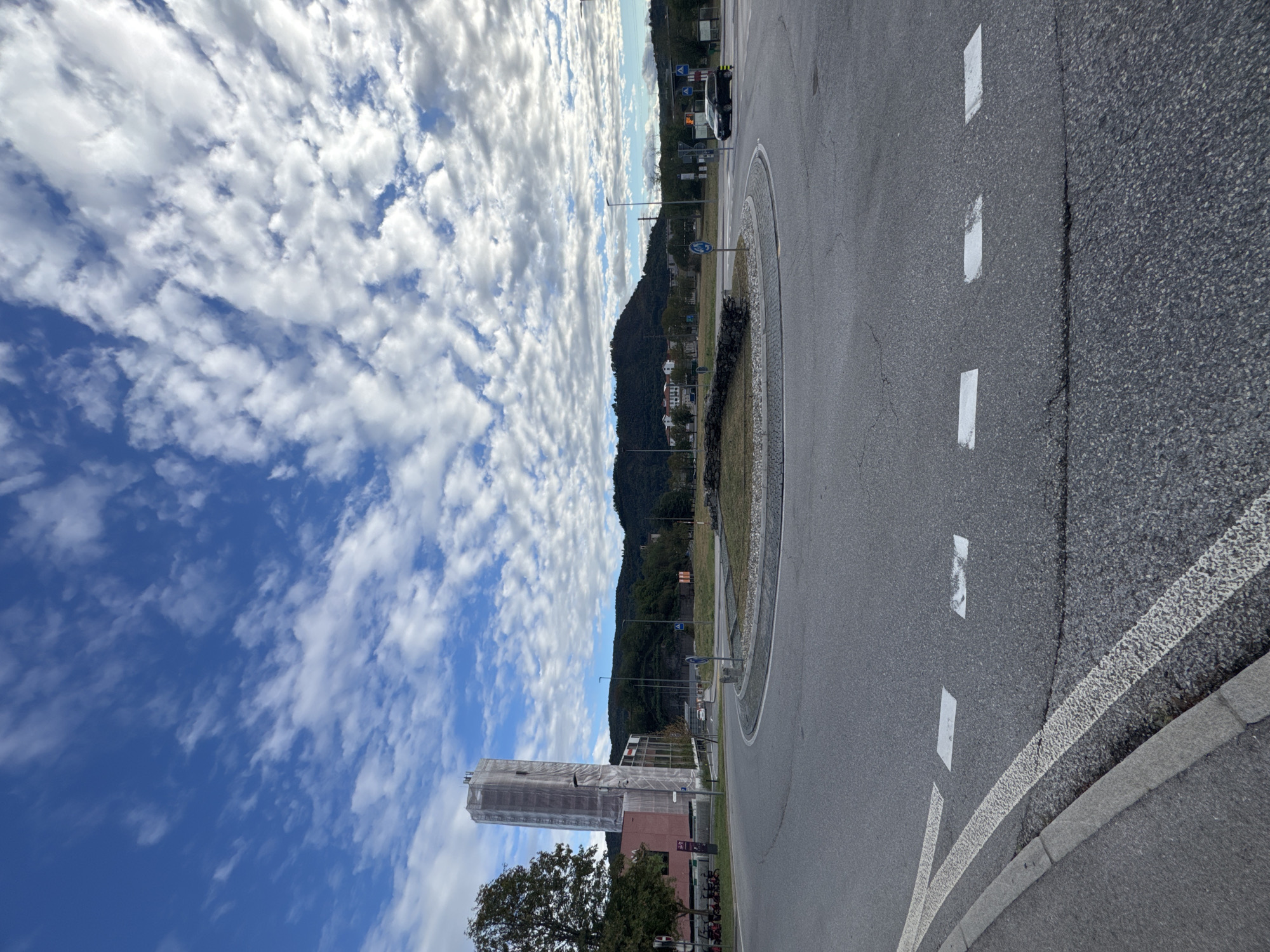 This is a wide-angle, eye-level, outdoor photograph taken in Morbio Inferiore, Switzerland. The image captures a roundabout on a paved road under a partly cloudy blue sky. In the foreground, the road surface displays white lane markings and a gray concrete curb.
The roundabout itself features a central island with low, textured stone walls and sparse, dry vegetation. A black car is visible in the distance, proceeding around the roundabout. To the left of the roundabout, a multi-story building is partially obscured by scaffolding and white sheeting, suggesting ongoing construction or renovation. Adjacent to this building, a row of red parked bicycles is visible.
In the midground and background, rolling hills covered in dense green trees rise behind a cluster of buildings with red-tiled roofs. The sky is a vibrant blue, dotted with numerous fluffy white clouds that create a dynamic texture. The overall lighting suggests daytime, possibly late morning or early afternoon, with soft shadows. The scene appears to be an urban or suburban setting, likely a transportation hub given the presence of the roundabout and the commercial or residential buildings.