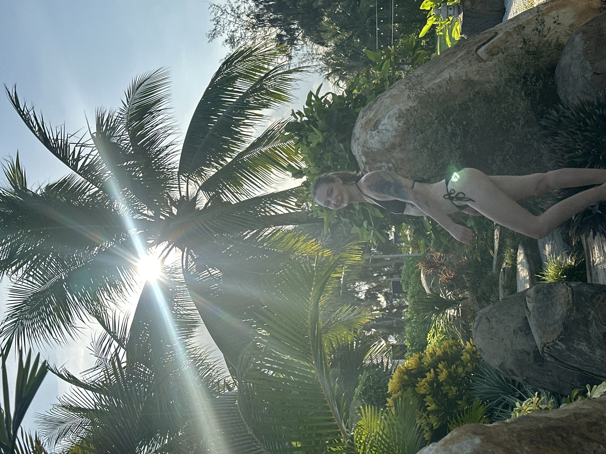 The media file depicts a woman in a bikini in a lush, tropical outdoor setting. She is positioned on the right side of the frame, lying on large rocks surrounded by dense green foliage, including palm trees. The sun is shining brightly, casting rays through the palm fronds, suggesting it is daytime, likely late morning or early afternoon given the angle of the sun. The woman appears relaxed and is smiling, indicating a pleasant and casual atmosphere. The environment suggests a location with a warm climate, such as Đà Nẵng, Vietnam, with its characteristic tropical vegetation and rocky outcrops often found near coastal areas or resorts. The overall impression is one of leisure and enjoyment in a beautiful, sunlit natural environment.