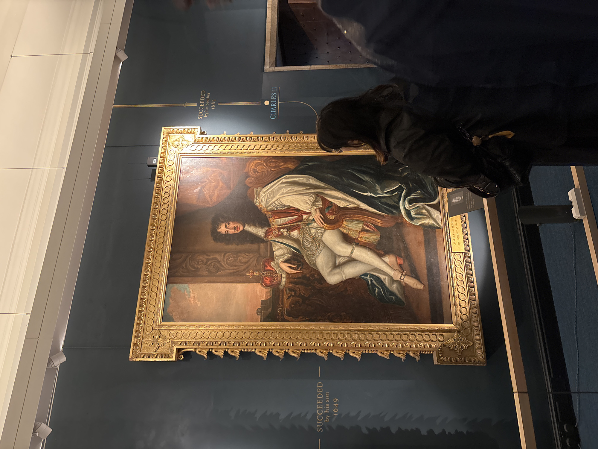 The image shows a vertical, eye-level view of an art gallery in the city of Edinburgh, United Kingdom. A framed portrait of Charles II hangs on a dark blue wall, accompanied by text panels indicating his predecessors and successors. A person, appearing to be a woman in a dark jacket, stands in front of the artwork, observing it. The frame of the painting is ornate and gold-colored, with intricate detailing. The gallery is well-lit, with the painting and text panels illuminated. A white wall with vertical partitions is visible on the left.