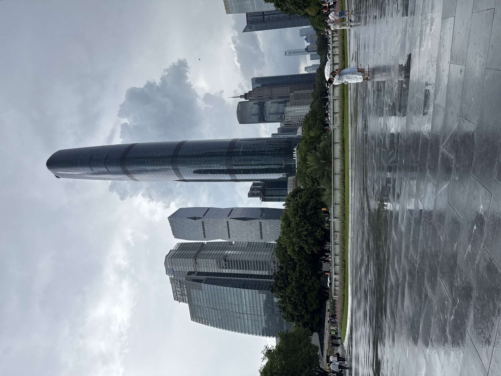 A vertical, eye-level, wide shot shows a cluster of modern skyscrapers under a cloudy sky in Tianhe District, China. In the foreground, a wet, dark, stone pavement reflects the overcast sky and the distant buildings. Several people are visible walking on the pavement and a grassy area behind it. To the left of the frame, a paved walkway curves away, with a line of trees and more people walking. A low wall with decorative plantings separates the walkway from a wider green space. The most prominent skyscraper, a tall, cylindrical building with a dark facade, stands in the center of the image, while other geometric, glass-fronted buildings are clustered around it. The sky is filled with heavy, gray clouds, suggesting recent or impending rain, which is evidenced by the wet pavement.