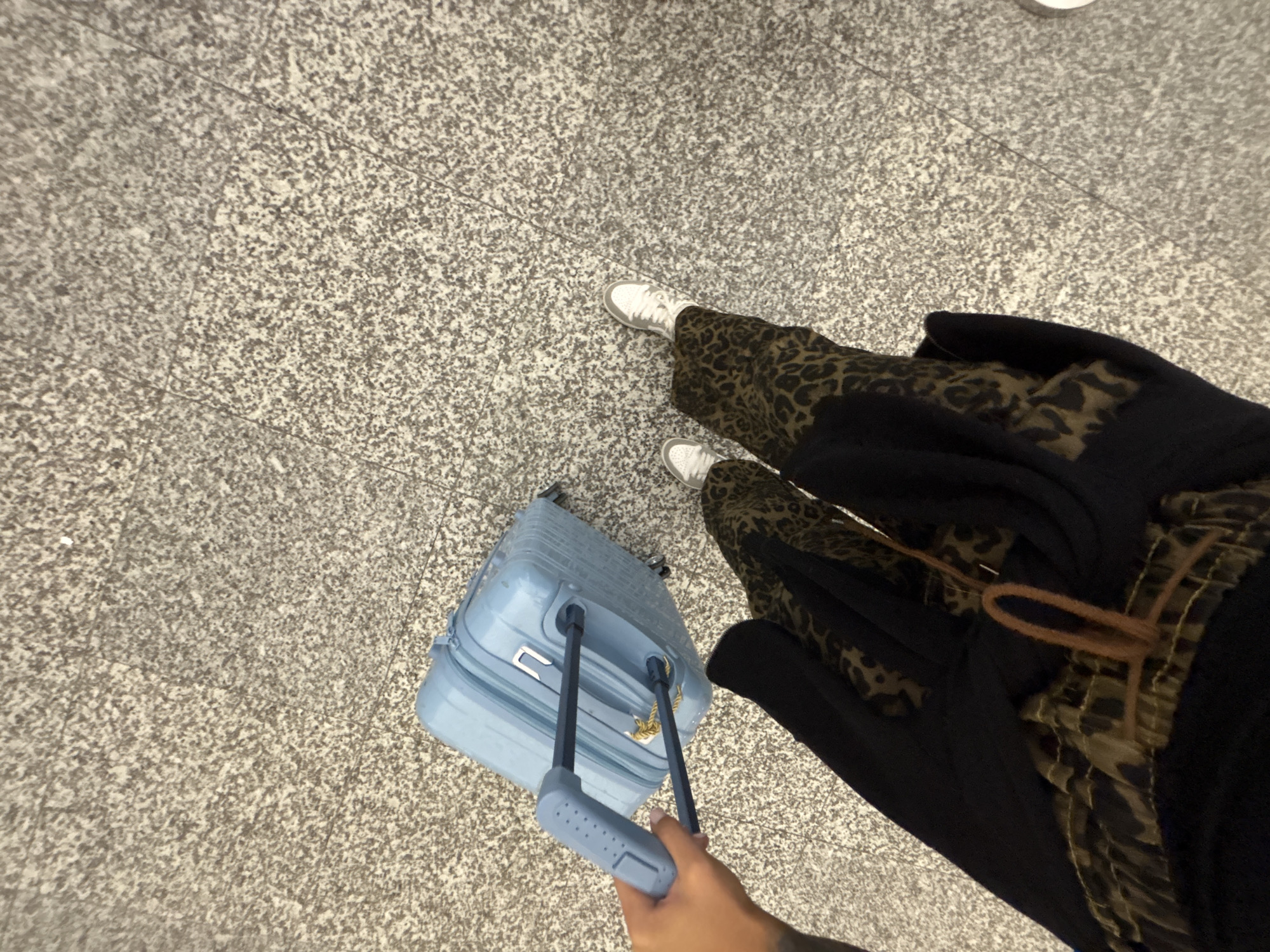 A person stands on a speckled gray floor, possibly granite or terrazzo, in an indoor location, likely an airport. The person is wearing white sneakers, animal-print pants, and a long black coat. They are holding the handle of a light blue suitcase. The background is out of focus, but appears to be a similar speckled gray floor. The location is unspecified in Kuta, Indonesia.