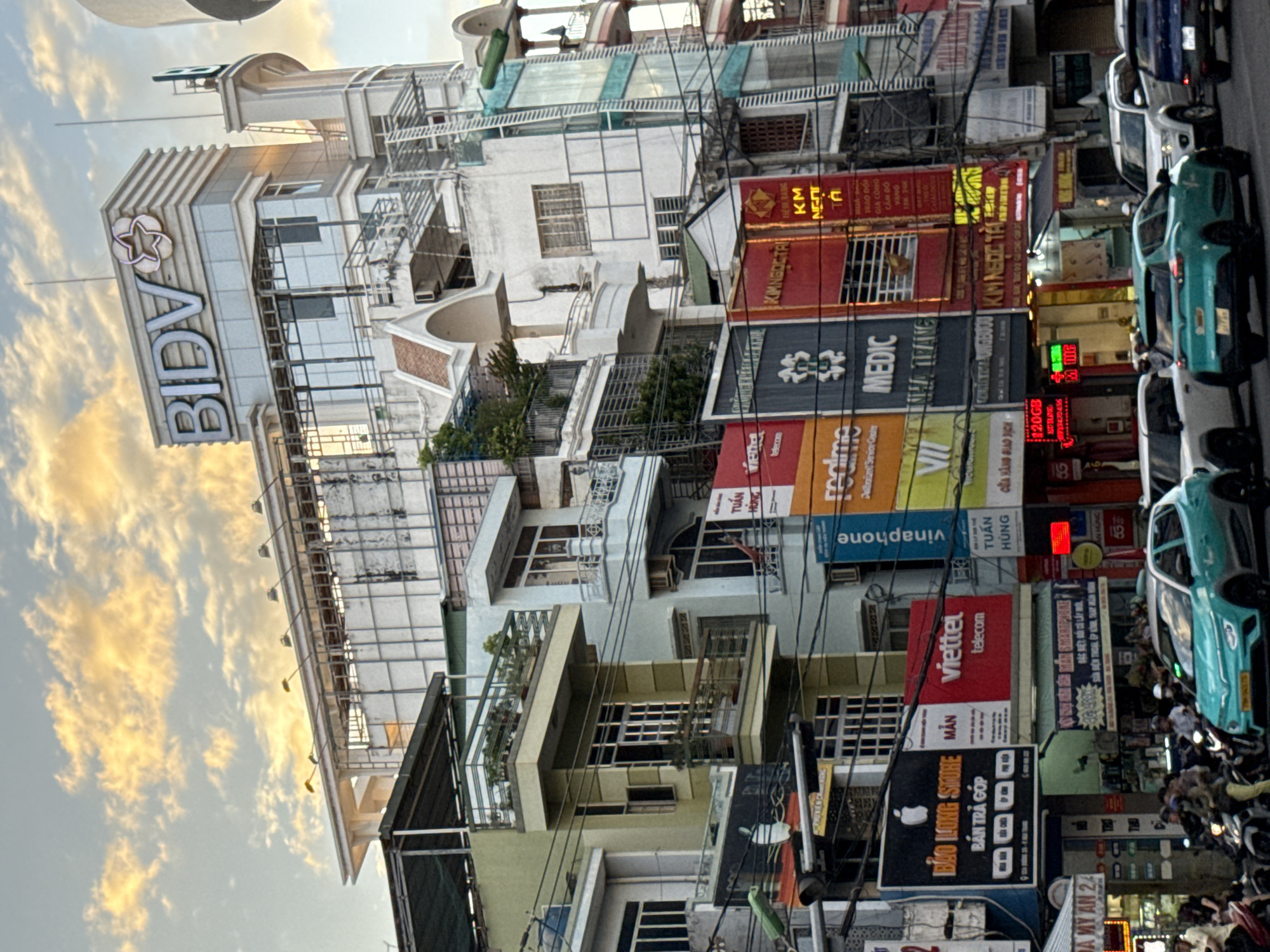 This is a medium-angle, slightly elevated shot of a busy street in Nha Trang, Vietnam, capturing a section of buildings with various shops and advertisements, along with traffic and pedestrians. The architecture features a mix of modern and older styles, with several multi-story buildings lined up. The sky above is partly cloudy with warm, golden light, suggesting either early morning or late afternoon.
Dominating the top of one building is a large sign for "BIDV," a Vietnamese bank, with its logo. Below this, the buildings display a multitude of colorful signs and advertisements, some in Vietnamese and some with English words. Notable advertisements include "realme," "VIETTEL telecom," "MEDIC," and "vinaphone." One prominent sign shows the Apple logo with "BAO LONG STORE."
On the street level, there is moderate traffic, with several cars and many motorcycles. A teal-colored electric car is prominently positioned in the foreground, and other vehicles, including white SUVs, are visible behind it. A few pedestrians are seen on the sidewalks and navigating through the traffic, some on motorcycles. The presence of power lines crisscrossing above the street adds to the urban, bustling atmosphere. The scene is vibrant and active, characteristic of a commercial street in a Vietnamese city.