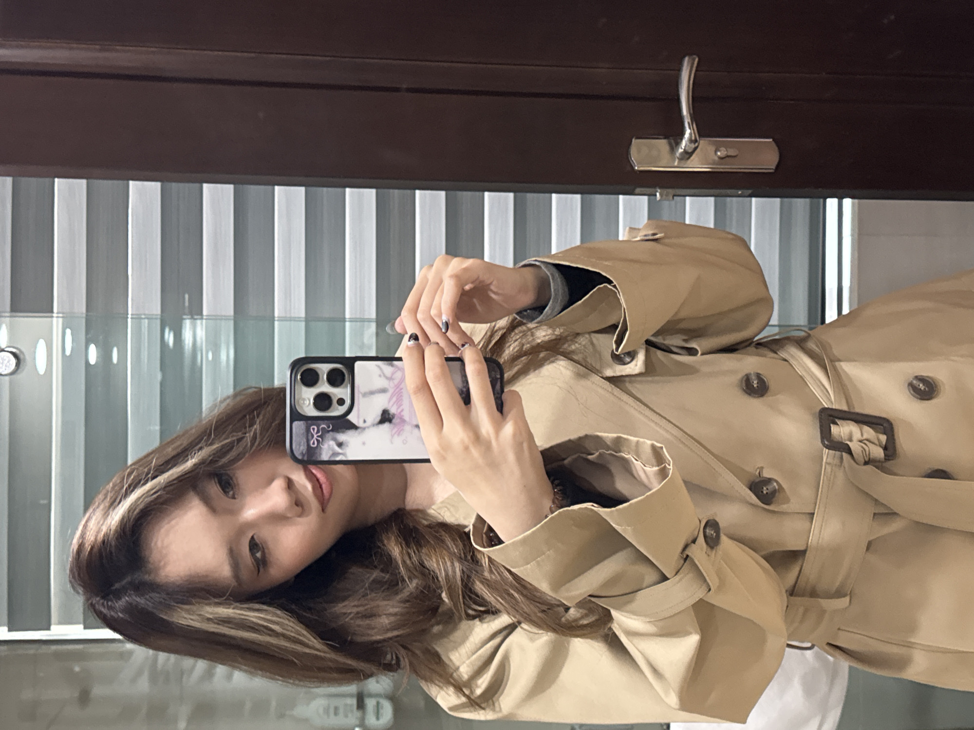 A woman with brown hair highlighted blonde is taking a selfie in front of a mirror. She is wearing a tan trench coat. The background features vertical blinds and a dark brown door with a silver hook. The reflection suggests the scene is indoors, likely a bathroom or closet area. The photograph does not provide sufficient information to confirm that the location is in the city of Lanzhou, China.