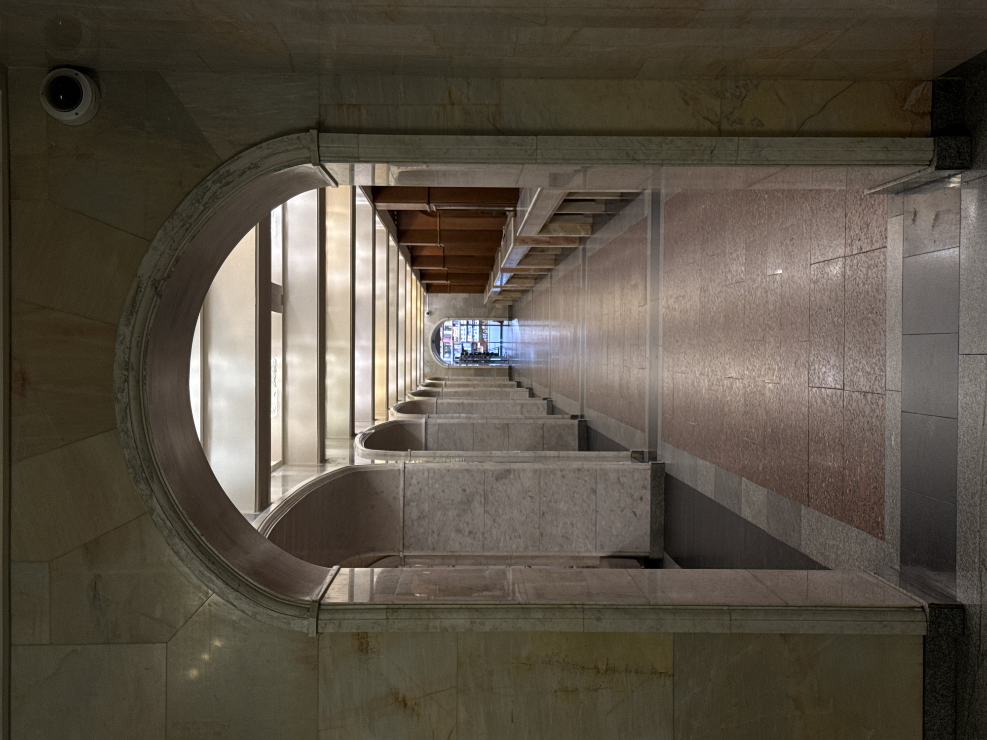 This is an interior shot of the Moscow State University Library in Moscow, Russia. The camera is positioned at a high angle looking down a corridor. The left side of the frame is dominated by a semicircular marble opening. Along the corridor, there are regularly spaced marble benches, each with a concave recess. The corridor floor is a patterned combination of gray and reddish-brown tiles. At the end of the corridor, a brighter area suggests an exit or opening with people visible. A security camera is visible in the upper left corner. The overall aesthetic is that of a grand, state-funded building.