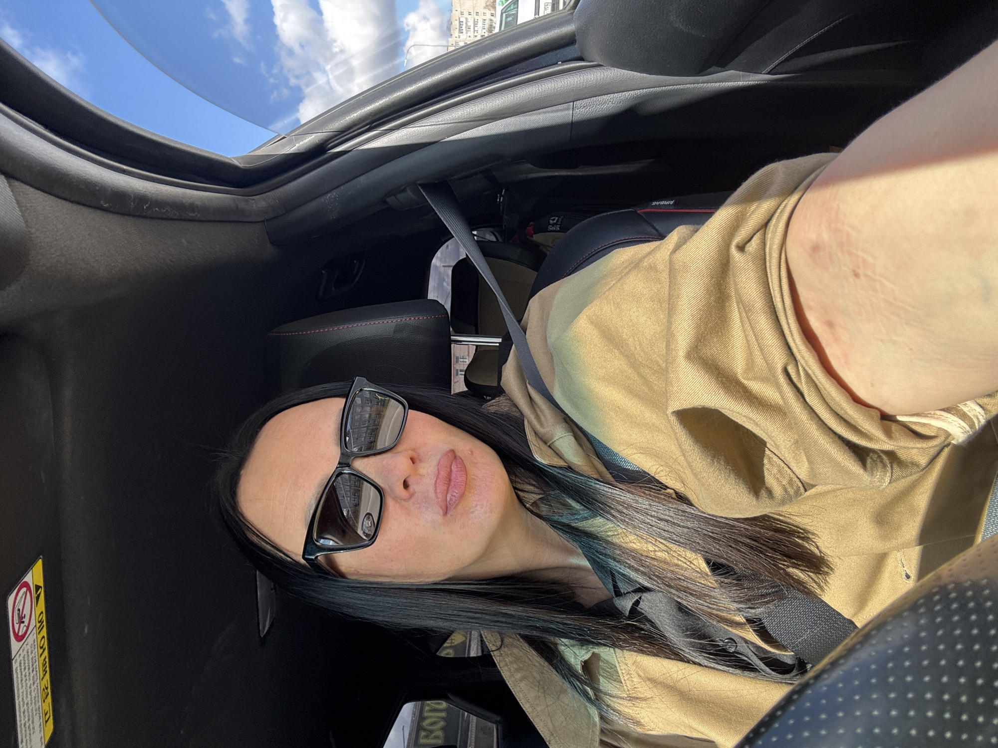 A person is captured in a selfie-style shot, sitting in what appears to be the driver's or passenger's seat of a car. The person has long, dark hair, some strands of which show a teal or blue tint. They are wearing large, dark-framed sunglasses that reflect a building structure. Their lips appear to have a light pink or nude color. They are dressed in a tan or olive-colored jacket or shirt, possibly a hooded garment, and are secured by a black seatbelt.
The setting is inside a car on a sunny day. The car interior features dark seats with red stitching and a black dashboard or door panel. The vehicle has an open sunroof or convertible top, revealing a bright blue sky with scattered white clouds. Sunlight casts strong shadows within the car.
Through the windows, distant buildings are visible, suggesting an urban or suburban environment. One of the cropped images shows a taller building with multiple windows. The sun is shining directly, creating a bright and clear atmosphere.
On the left side of the frame, a yellow warning label or sticker is partially visible, oriented vertically. Although most of the text is illegible due to the angle and partial view, the Japanese characters "注意" (Chūi - caution/warning) are somewhat discernible. In the reflection of the sunglasses, building windows are visible. Another reflection, possibly in the lower part of the image, shows faint Cyrillic text, potentially "БОГО" (BOGO), but this is very indistinct. The provided location context is Chaikovsky, Russia.