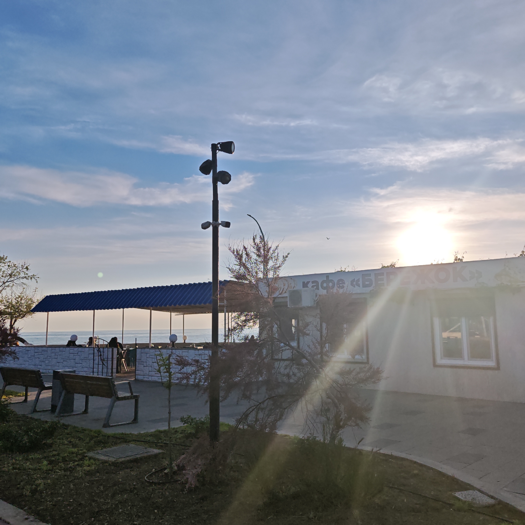 The image depicts a scene at a cafe near the sea in городское поселение Щелкино, Ukraine. The cafe, named "БЕРЕЖОК," is a modest, single-story building. Sunlight streams past the building, creating glare. A blue-roofed structure with supporting columns stands adjacent to the cafe, providing covered seating with a view of the sea. People are seated beneath this structure. In the foreground, there are two wooden benches with metal frames on a grassy area next to a paved walkway. A streetlamp with three lights stands nearby. A small tree is also visible near the benches. The sky is partially cloudy, with the sun visible.