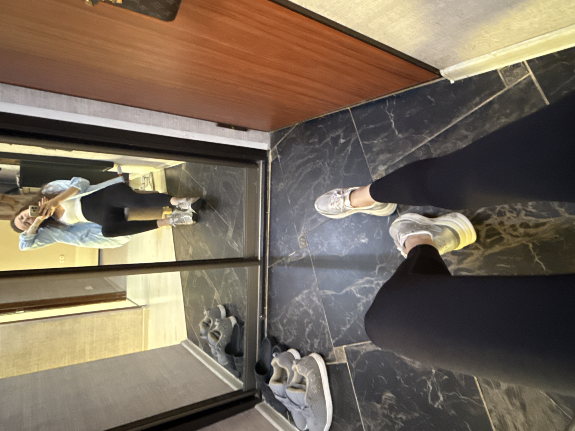 This image captures an indoor scene, likely a hallway or entryway in Moscow, Russia, taken from a high-angle perspective and rotated ninety degrees clockwise. The primary subject is a person taking a mirror selfie, reflected in a large, framed mirror. The person in the reflection is seen from the chest down, wearing black leggings, a white tank top, and a light blue long-sleeved shirt or jacket unbuttoned. They are holding a light-colored smartphone to take the picture, and their light-colored sneakers are visible.
In the immediate foreground, the feet and lower legs of the person taking the photograph are visible, also wearing black leggings and chunky, light-colored sneakers with reflective accents. These feet are positioned on a dark, glossy floor with a grey-white marble pattern. The mirror itself has a thin dark frame. Adjacent to the mirror, on the right side of the image, is a section of dark, wood-paneled wall or a door, and above that, a lighter-colored wall or ceiling. On the floor beside the mirror, another pair of grey slip-on shoes with white soles are neatly placed. The overall lighting suggests an indoor setting during daytime or a well-lit interior. No visible text is present in the image, and the emotion conveyed is neutral, typical of a casual selfie.