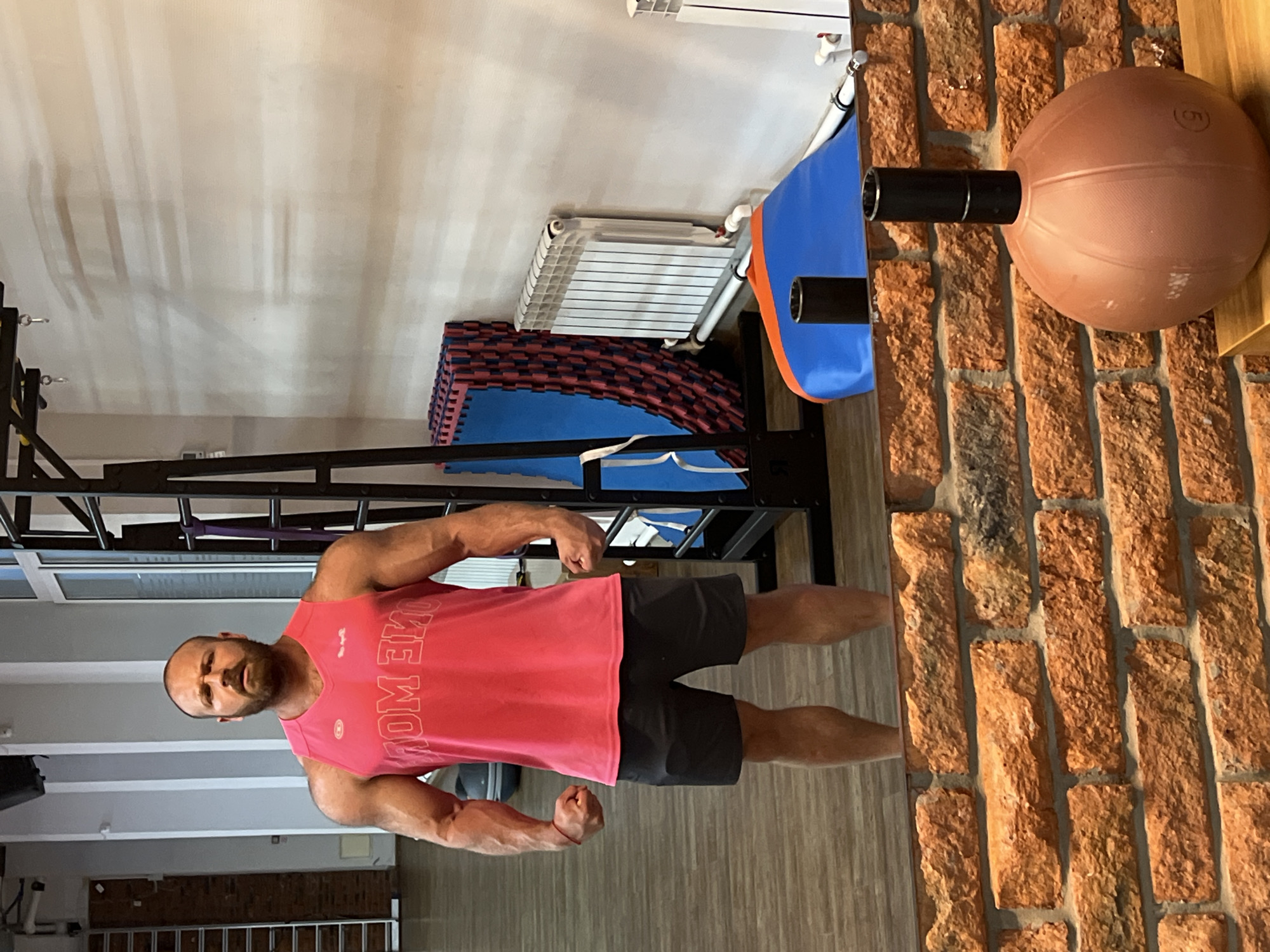 A man with a bald head and beard, wearing a pink sleeveless t-shirt and black shorts, poses in what appears to be a gym. He is flexing his arms, showcasing his muscular physique. In the background, there is a brick wall with a large brown ball mounted on it, possibly a medicine ball or a punching ball. Adjacent to the wall are some stacked mats, a radiator, and part of a workout structure. The lighting suggests it is daytime. The man's expression is serious and focused. The t-shirt has visible text on it, although it is partially obscured and reversed due to the angle of the photo, it appears to say "COME ON". The overall scene depicts a workout environment.