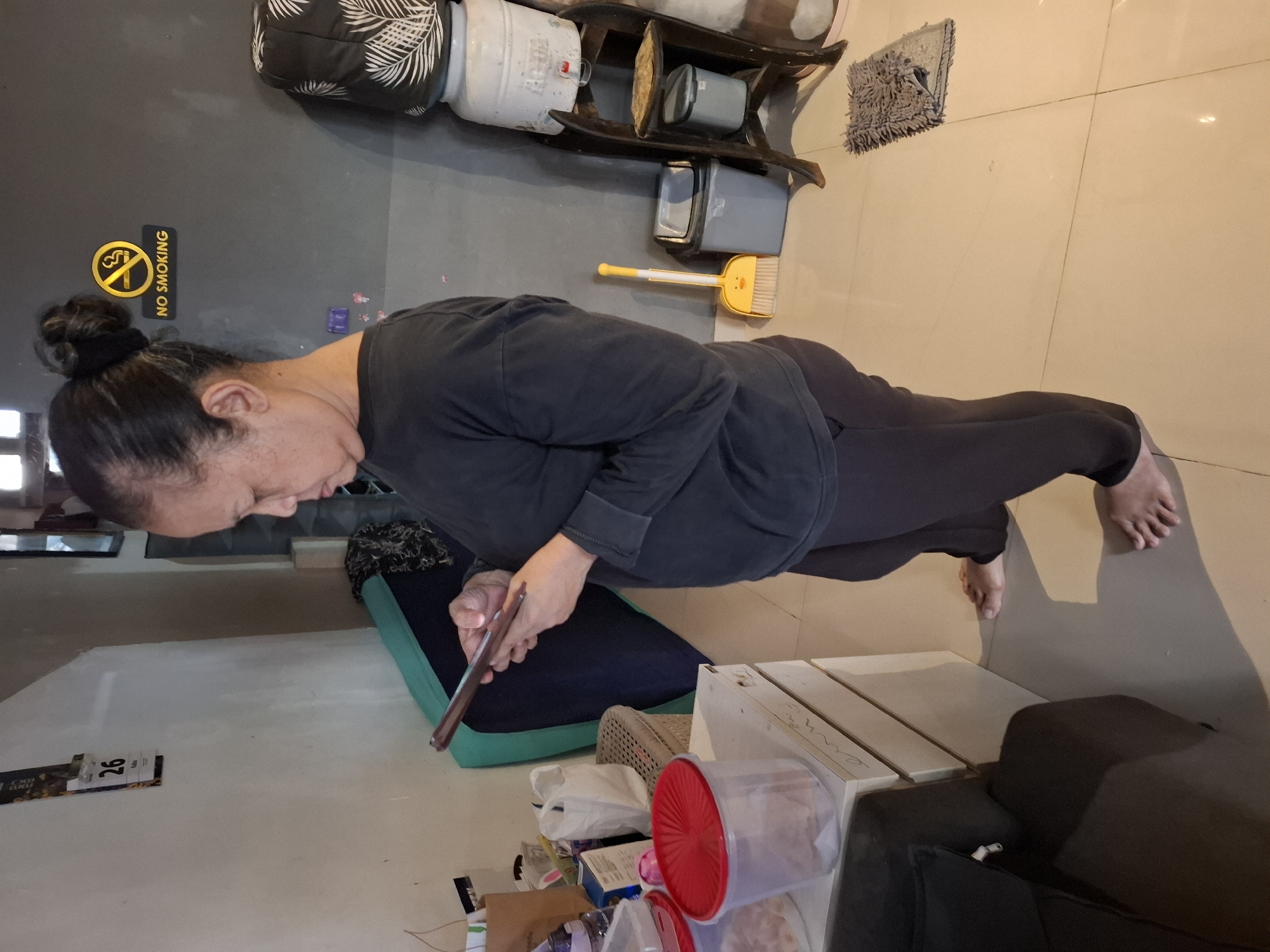 In the city of West Jakarta, Indonesia, a woman stands in a domestic setting, looking at her mobile phone. She has dark hair pulled up in a bun and is wearing a dark long-sleeved shirt and dark pants. The room appears to be a kitchen or utility area. To her left, a white cabinet is visible, holding containers, one of which has a red lid. Behind her, a dark blue sofa or mattress is present. To her right, on a grey wall, a "No Smoking" sign is displayed. Further to the right, a water dispenser sits atop a wooden shelf unit. On the floor, a yellow dustpan with a smiley face and a broom are placed next to a grey bin. The floor is tiled in a light beige color. A calendar with the number "26" is affixed to the wall on the far left.