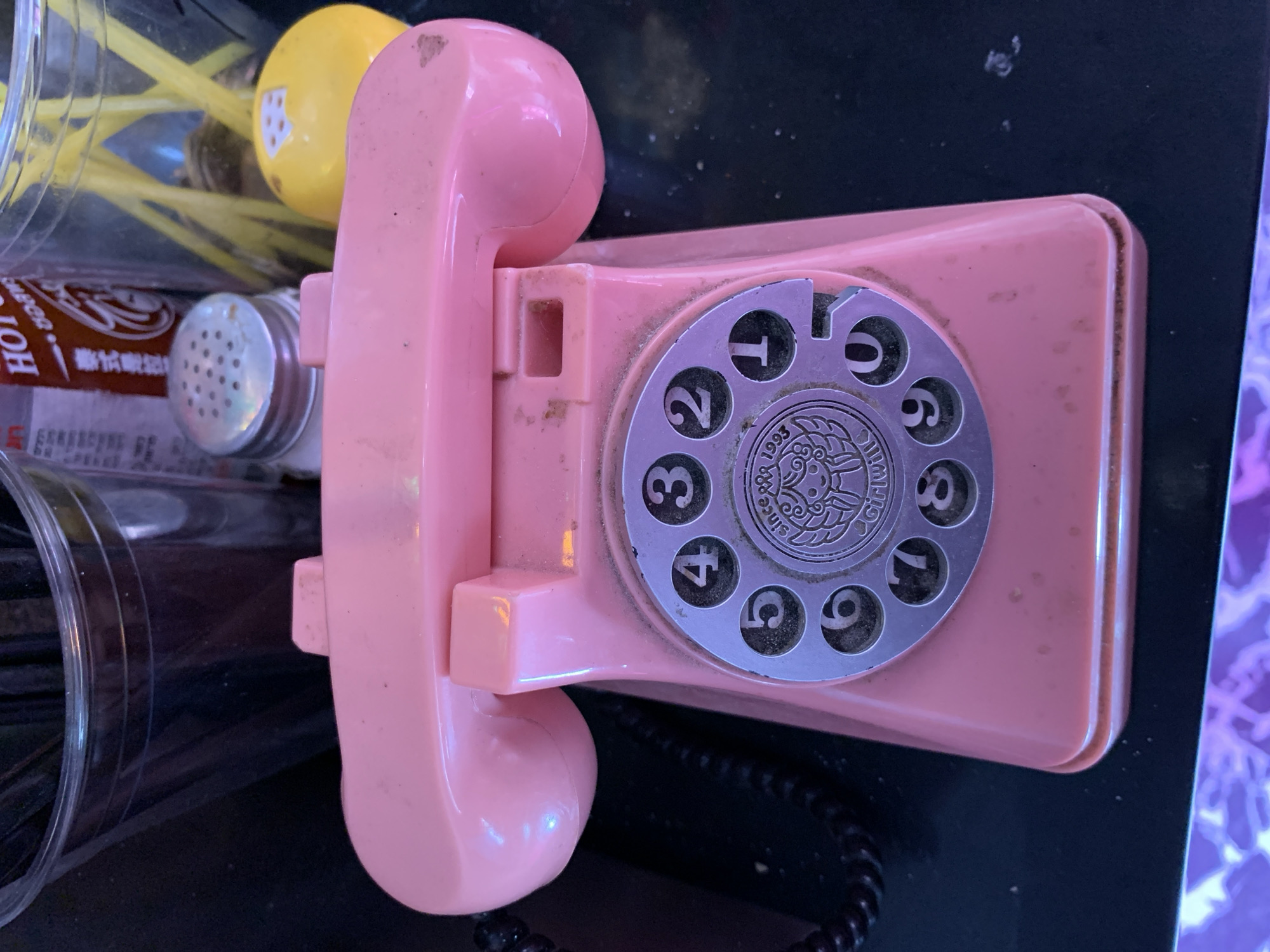 Here is a description of the image:
The image shows a pink rotary dial telephone sitting on a black surface, likely a table. The dial is silver with black numbers. The area surrounding the phone contains various objects, including a jar of hot sauce, a salt shaker, and a container holding what appear to be yellow and black straws. The scene is generally cluttered and may be located indoors. There is no information available to confirm if the setting is Tel-Aviv, Israel.