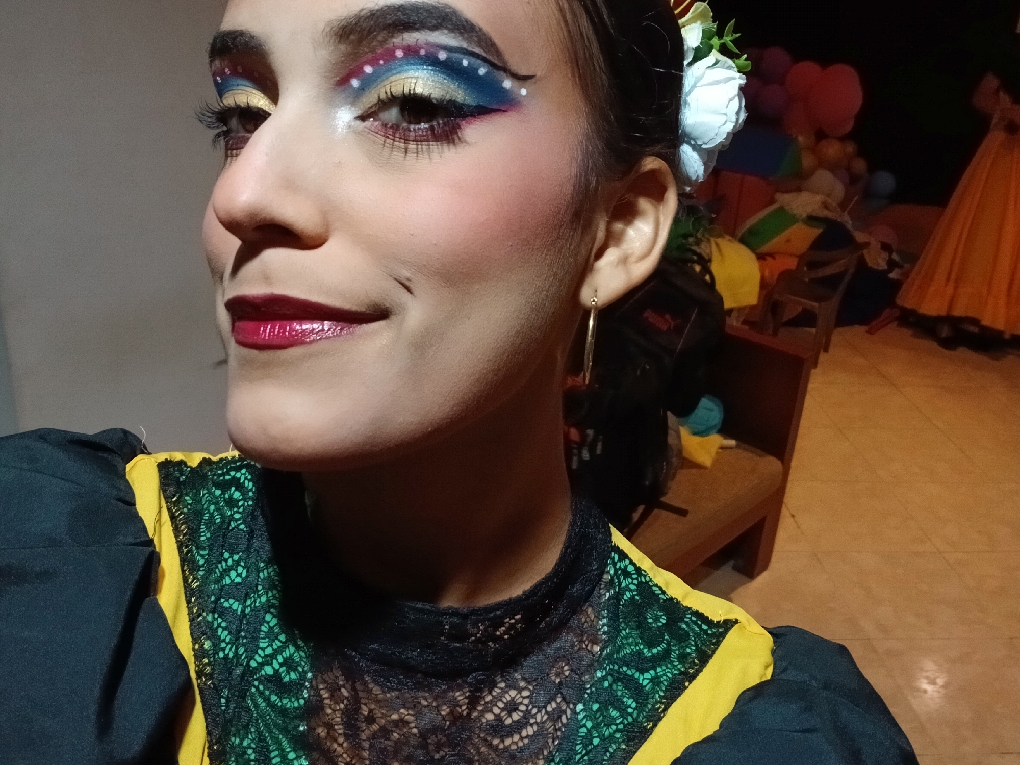 A close-up image shows a woman with elaborately decorated makeup, likely in a theatrical or performance context. The woman is wearing striking makeup including colorful eyeshadow, heavy eyeliner, and a bold red lipstick. She is wearing a dark-colored dress with yellow and black lace accents, and a white flower is tucked behind her ear. The photo was taken at a low angle.
Behind her, there are hints of a chaotic backstage area, including balloons, a wooden chair, and another person in a yellow dress. Although the prompt mentioned Playa El Agua, Venezuela, there is no concrete information in the picture to confirm the location.