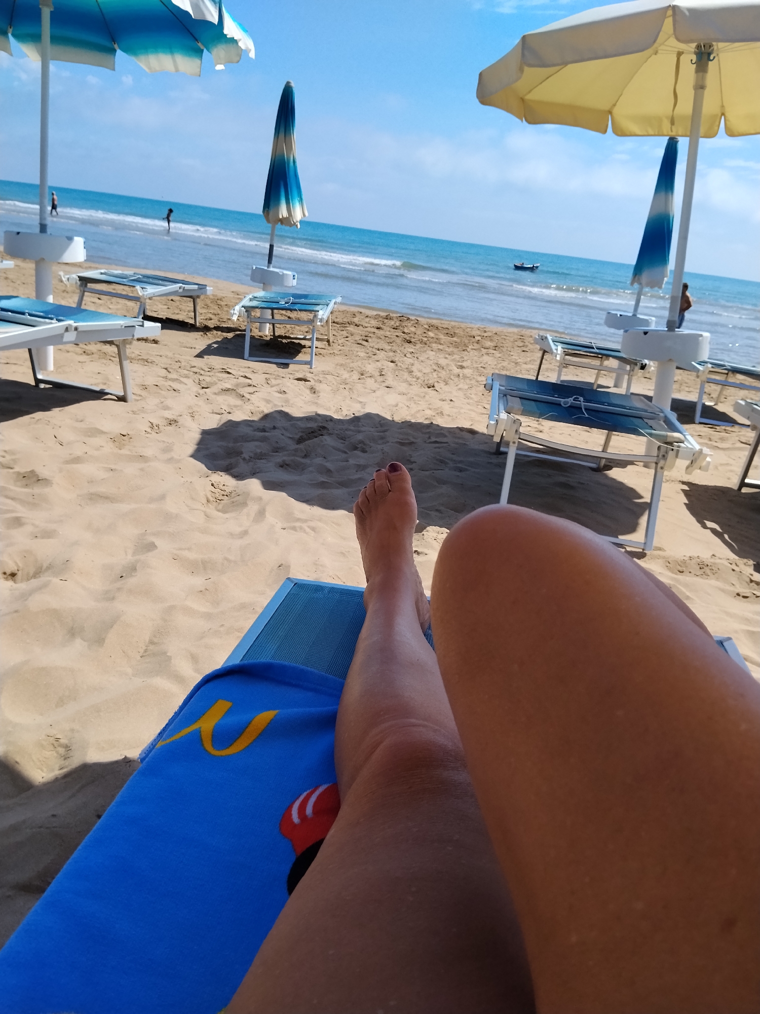 A first-person perspective shot captures a beach scene in Peschici, Italy. The foreground is dominated by a person's legs resting on a blue beach chair covered with a blue towel featuring a yellow curved symbol and a red and white circular image. The sandy beach stretches out behind, dotted with empty beach chairs and umbrellas. In the background, the calm blue sea meets the horizon under a clear sky, with a small boat visible on the water and a couple of distant figures walking near the shoreline. The overall impression is one of a relaxed day at the beach.