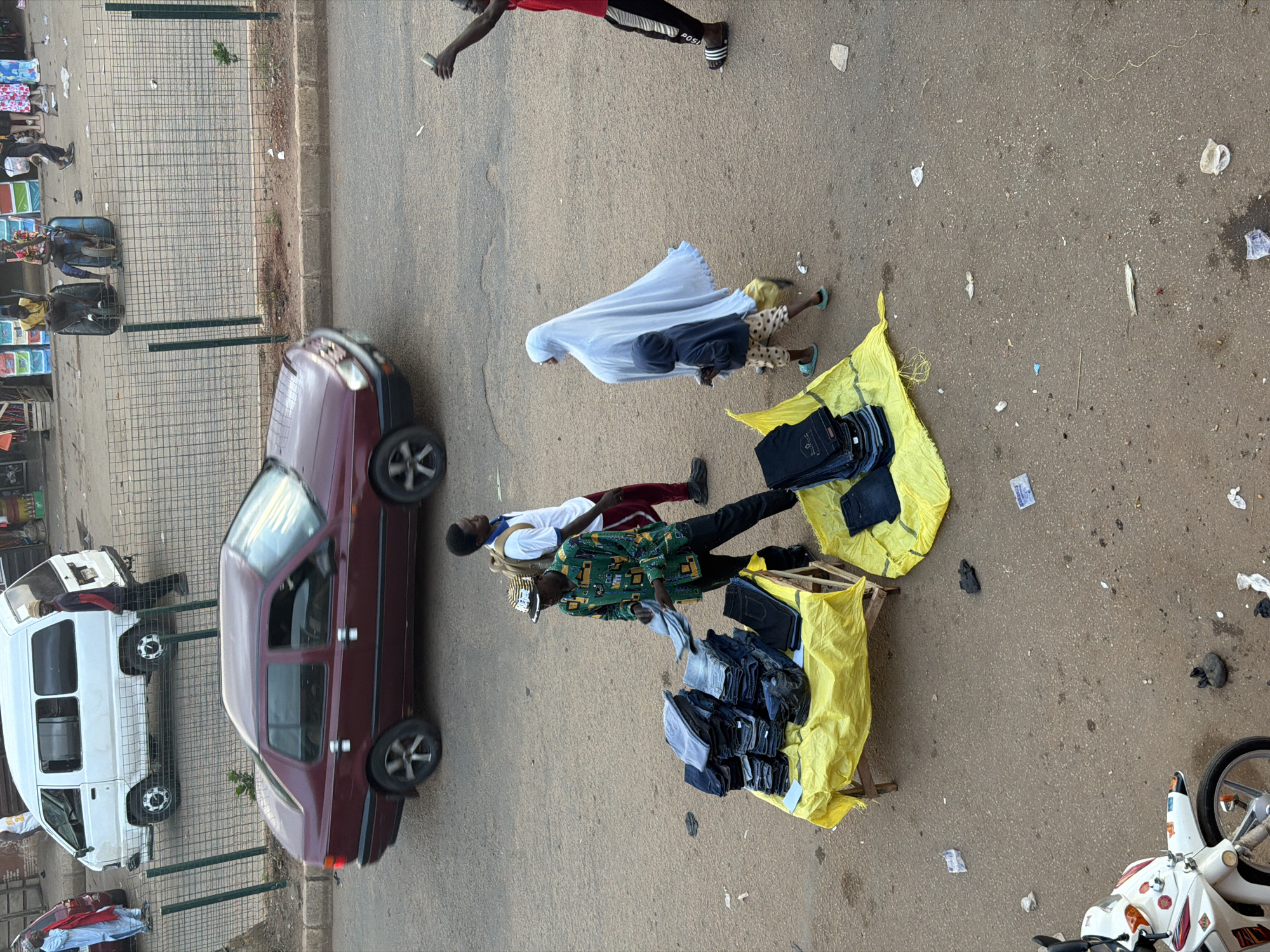 The image depicts a street scene in Bukuru, Nigeria. In the foreground, a street vendor is displaying stacks of folded jeans on a yellow tarp laid over a wooden stand. The vendor, wearing a patterned shirt and a hat, is interacting with a customer. Behind the vendor, a person wearing a white shirt and red pants walks by, carrying a backpack. To the right of the vendor, a woman in a light blue hijab and a child in a blue hooded jacket walk past. Further in the background, a dark red sedan drives by. On the left side of the image, a white van is parked. The street appears to be paved with asphalt, and there is a metal fence in the background separating the street from what seems to be a market or roadside stalls, where other people are visible. Scattered debris, including pieces of paper and plastic, is present on the ground. A white motorcycle is parked in the lower left corner of the image.