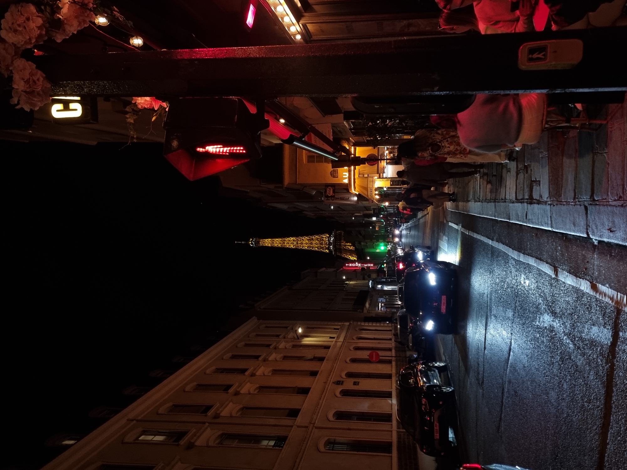 This nighttime street scene in Paris, France, features the Eiffel Tower illuminated in the distance. The view is looking straight down a street with buildings lining each side. Cars are parked on the right side of the street, their headlights reflecting off the wet pavement. Pedestrians are walking on the sidewalk on the right. Above the sidewalk is a structure with lights, signage, and floral decorations, partially obscuring the view. A traffic light is visible in the upper center of the image, showing a red light.