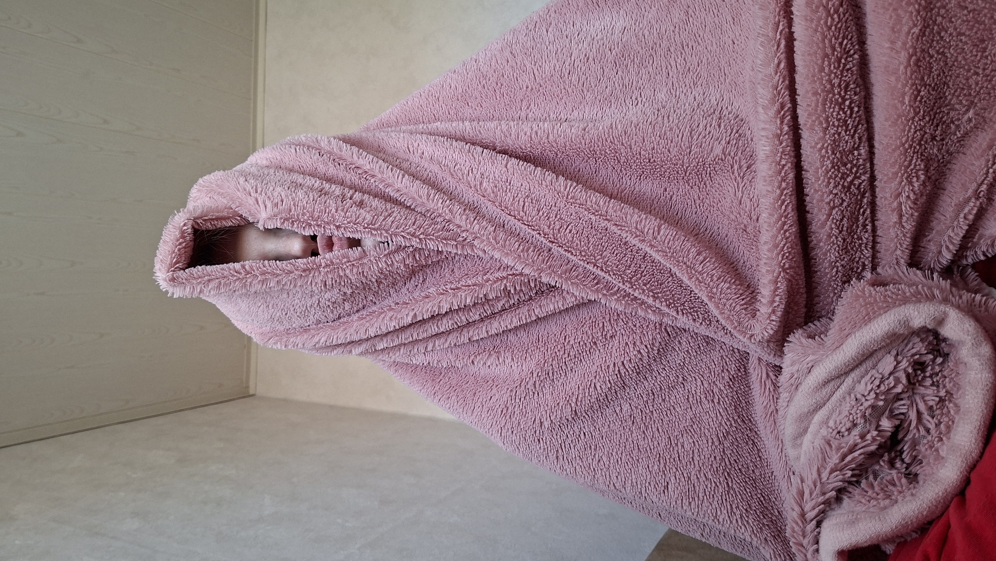 A person is enveloped in a pink, fluffy blanket or robe, obscuring most of their body except for a glimpse of their face and possibly their teeth visible through a small opening. The background shows a wall and floor, suggesting the scene takes place indoors, possibly in a residential room. The room appears to be in the city of Большой Букор, Russia.