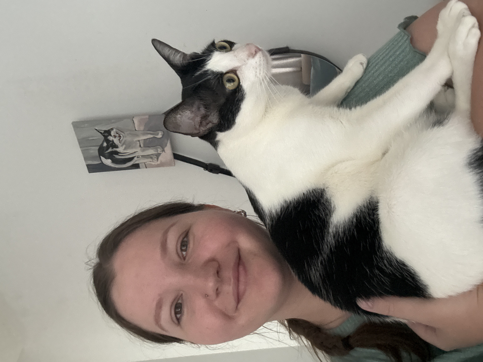 A woman is holding a black and white cat. A painting of the same cat is on the wall behind them.