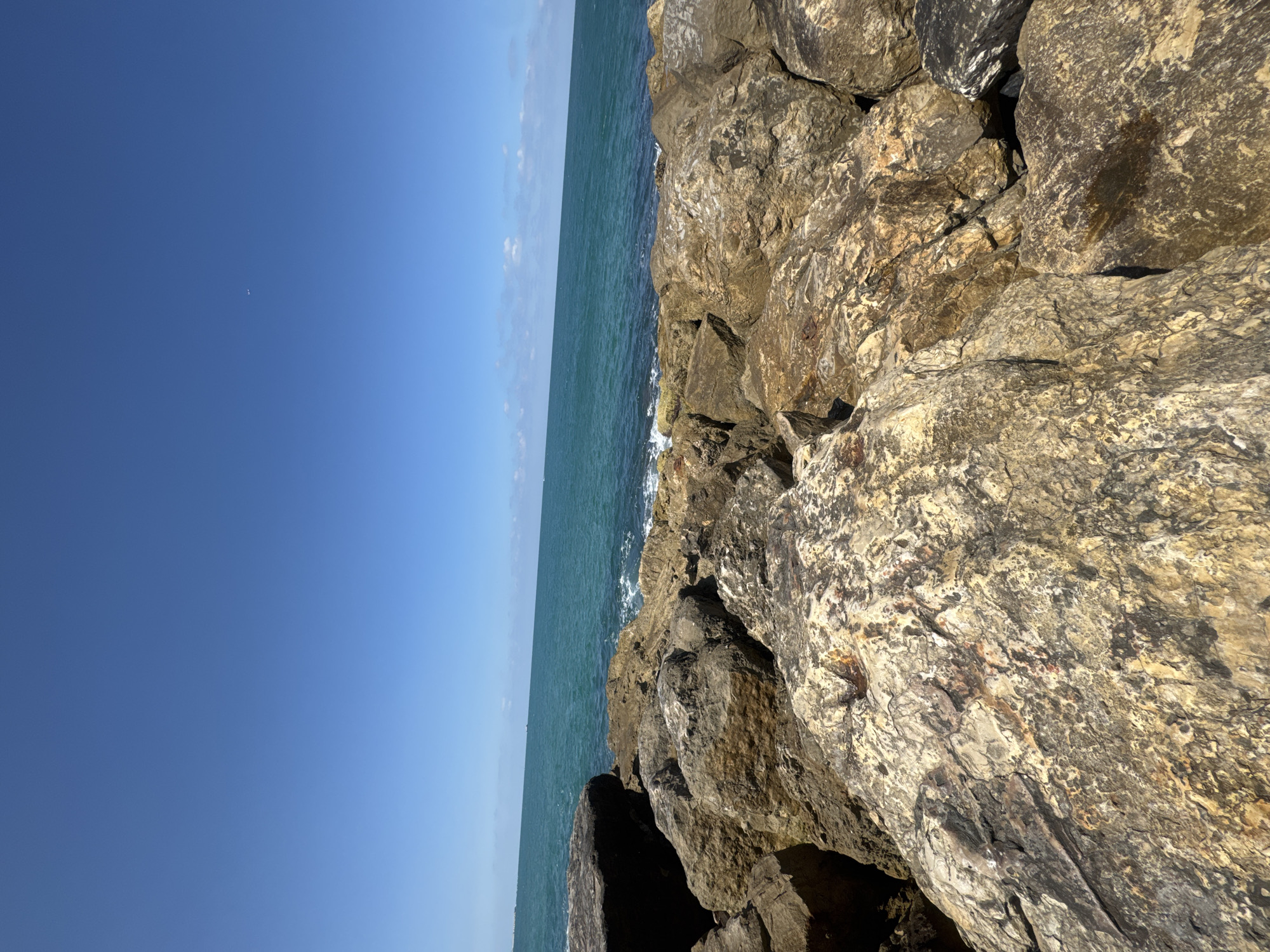 The image shows a rocky breakwater along the coast of Tel Aviv, Israel. The breakwater is constructed of large, unevenly shaped, tan and gray rocks. The Mediterranean Sea is visible beyond the rocks, with gentle waves washing against the structure. A clear blue sky is visible above the sea. The horizon is a straight line separating the sea and sky. There are no people or other objects visible in the image.