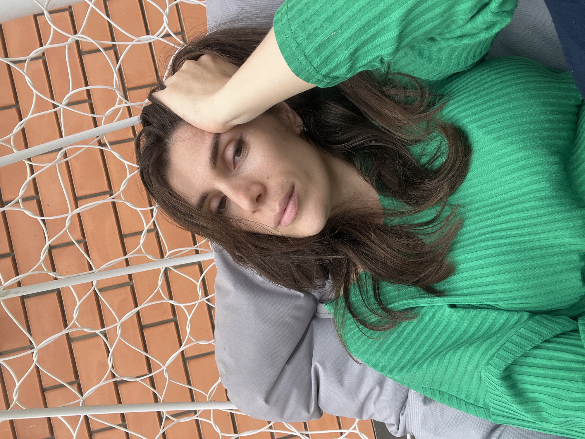 A young woman lies on a white wire hammock in what appears to be an outdoor setting. She is reclining on a grey pillow. Above the hammock, there is a brick wall. The woman is wearing a green ribbed shirt. She has brown hair and is resting her hand on her forehead. The location in the city of Kaspiysk, Russia is not discernible from the image.