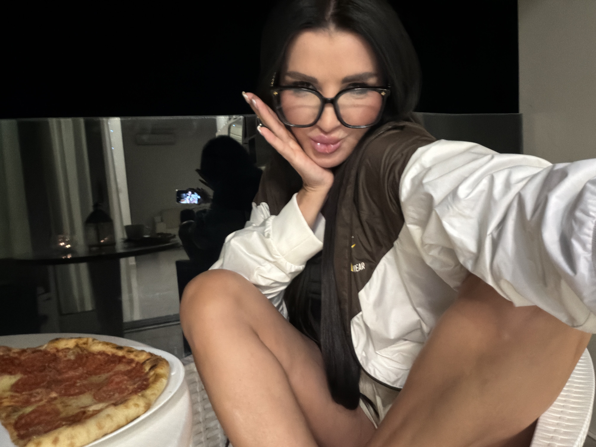 A woman takes a selfie at night, likely on a balcony in Boreti, Montenegro. She has long dark hair, wears large black-rimmed glasses, a white jacket or shirt, and a brown vest. She poses playfully, pouting and resting her hand against her cheek. A plate with a pepperoni pizza is visible in the foreground to her left. In the reflection on the glass behind her, a dark silhouette of a person holding a phone is visible. The background outside the balcony is dark, indicating nighttime. The word "WEAR" is partially visible on her brown vest. Her legs are crossed, suggesting she is seated in a relaxed position.