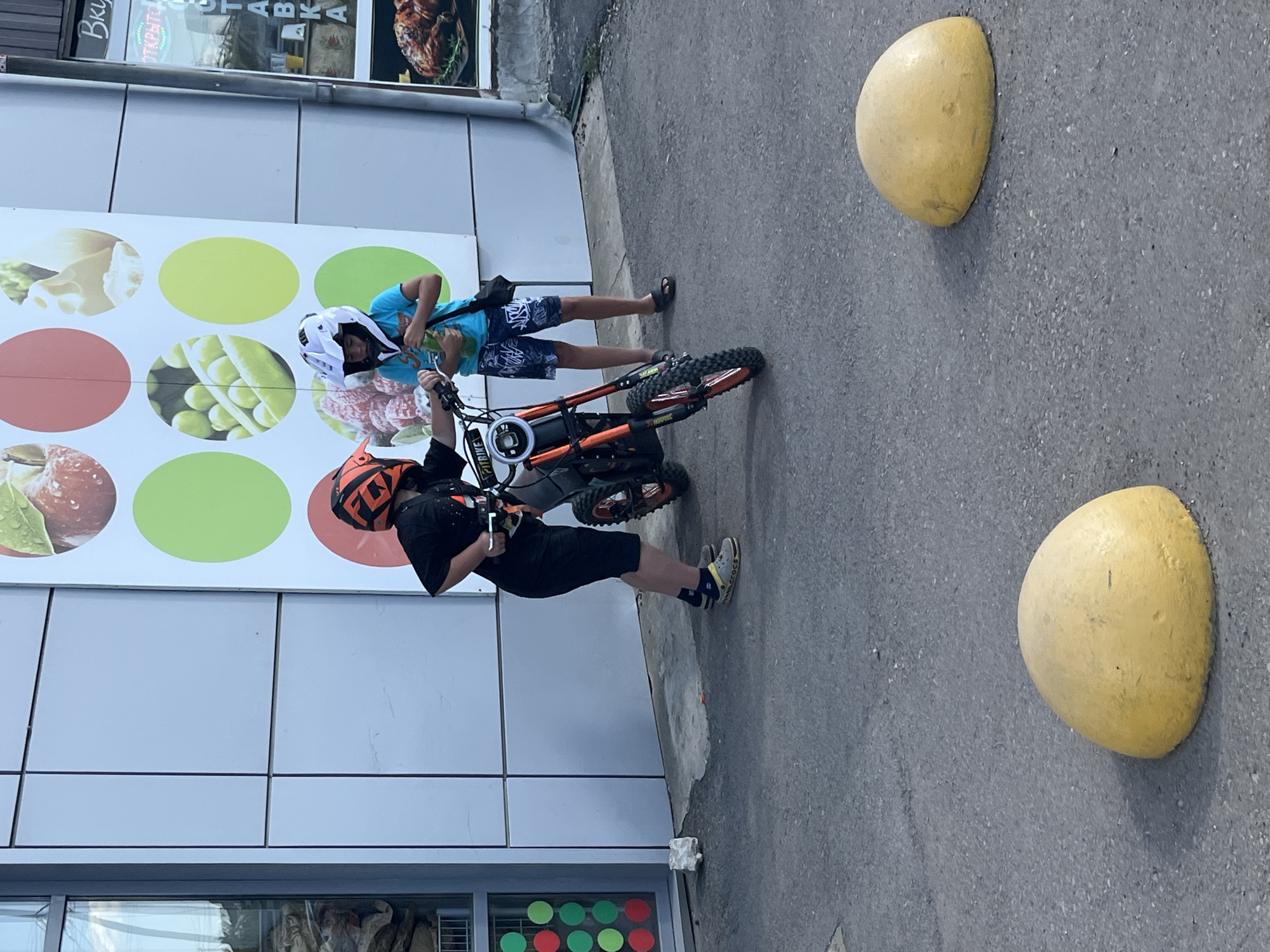 The image shows two young boys standing near a small, orange-colored dirt bike parked on an asphalt surface. The boy on the left, wearing a black t-shirt, dark shorts, and an orange helmet, is reaching for the handlebars of the bike. The boy on the right, wearing a blue t-shirt, patterned shorts, and a white helmet, is holding a green bottle and looking at the bike. They are standing in front of a building with a light-colored, paneled facade and a large mural depicting fruits and vegetables. Yellow, semi-spherical bollards are visible in the foreground. The scene appears to be outdoors, likely in a public area like a parking lot or sidewalk.