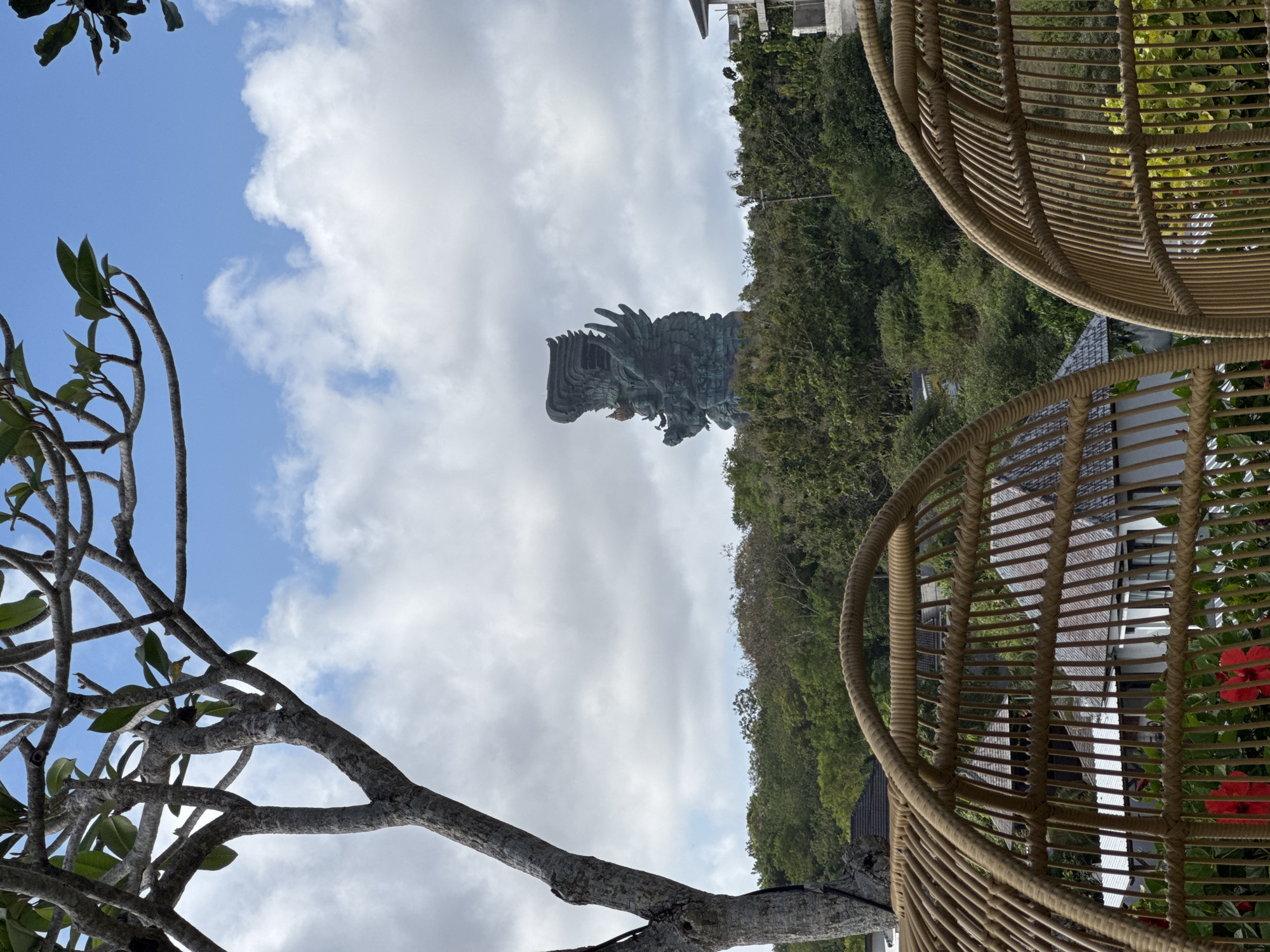 The image shows the Garuda Wisnu Kencana statue in Kuta Selatan, Indonesia, partially obscured by trees and clouds. The statue is centrally positioned against a sky with scattered clouds. Two rattan chairs frame the right side of the image, with additional greenery visible behind them. A tree branch with sparse leaves is visible on the left side. The perspective is from a slightly low angle, looking upwards toward the statue.
