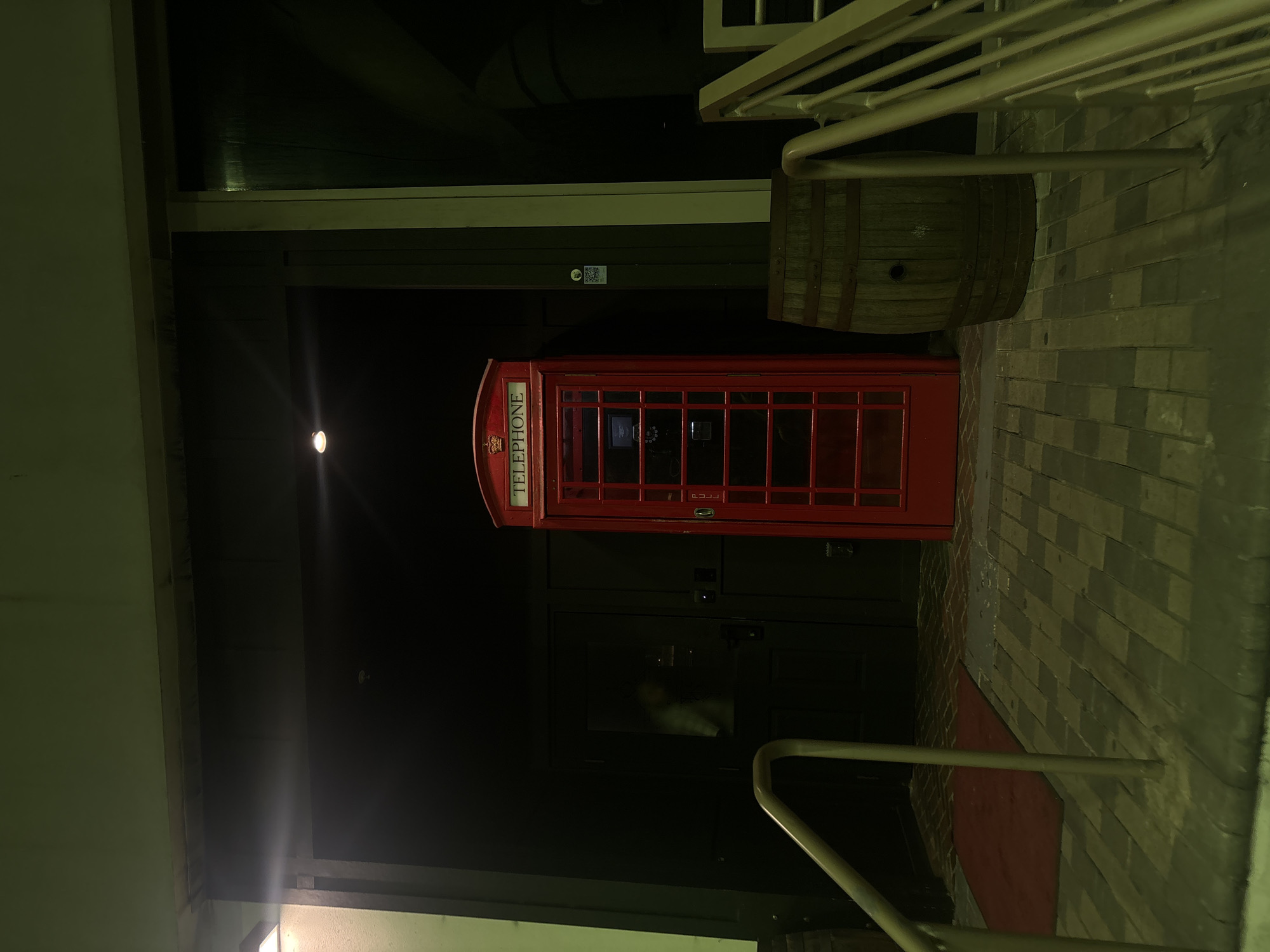 This image captures a dimly lit exterior scene at night, depicting the entrance to a building in Miami, United States of America. Central to the frame is a classic red British telephone booth, prominently displaying "TELEPHONE" and a crown emblem on its upper panel. The booth features glass panes revealing a rotary telephone instrument inside.
The telephone booth stands directly in front of dark, recessed double doors, which appear to be the primary entrance to the building. Through the glass panel of the left door, a faint, translucent figure of a person, seemingly wearing a light-colored top, is visible. Above the doors, a single recessed light fixture emits a warm glow, creating a starburst effect.
To the right of the telephone booth, an upright wooden barrel with a bung hole is positioned. Adjacent to the barrel and extending towards the right edge of the frame are metal handrails, indicating the presence of a staircase ascending out of view. Another wooden barrel is partially visible on the far left of the image. A white sticker with a black QR code is affixed to the dark wall panel to the right of the telephone booth.
The ground in the foreground is paved, featuring a reddish brick pattern beneath a rectangular red doormat directly in front of the doors, transitioning to a pattern of dark rectangular blocks closer to the viewer. Metal handrails are also present on the left side of the frame, leading downwards towards the foreground. The overall lighting is low, with visible lens flares from bright light sources located off-frame to the left and directly above the entrance.