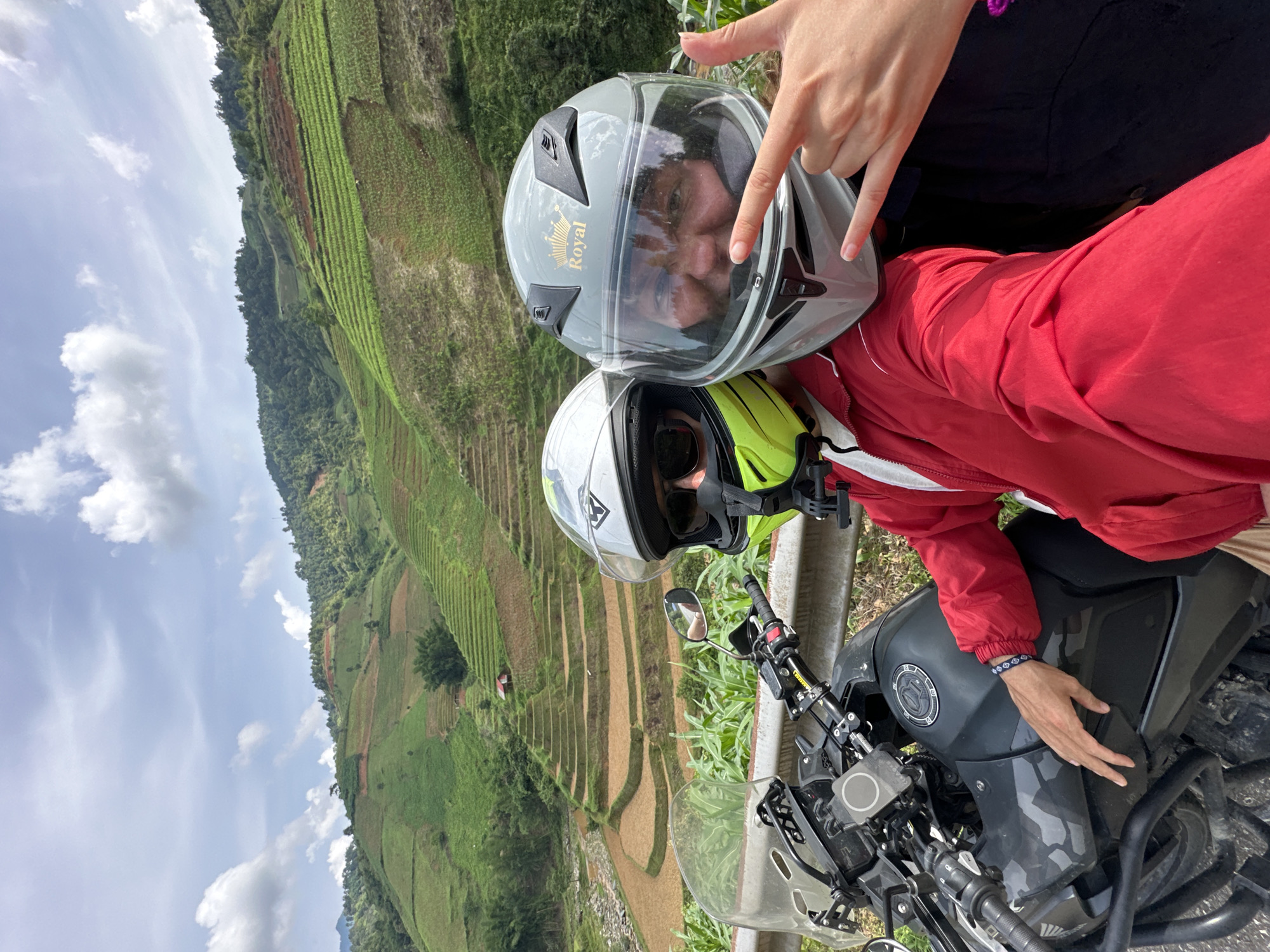 Two individuals on a motorcycle are captured in a bright, outdoor photo, likely taken during a stop on a scenic route in Khao Mang, Vietnam. The image, originally in portrait orientation, features a stunning landscape of terraced fields in the background.
In the foreground, two people are wearing motorcycle helmets. The person closer to the camera wears a grey full-face helmet with a clear visor, reflecting a face and showing a gold "Royal" crown logo. This person is making a "peace sign" gesture with their right hand. They are wearing a dark top. Behind and slightly to the right, the second person wears a white and neon yellow helmet, with a dark visor or sunglasses visible. This individual is dressed in a vibrant red long-sleeved jacket and has their right hand resting on the fuel tank of a dark-colored adventure-style motorcycle. A blue and white patterned bracelet is visible on their wrist.
The motorcycle, partially visible, features a windscreen and handlebars. Beyond the subjects and a metal guardrail, the landscape unfolds with breathtaking terraced fields, characteristic of rice cultivation in mountainous regions. These green fields step down the slopes, interspersed with darker green foliage and patches of brown earth. A blue sky with scattered white clouds completes the picturesque setting, indicating a clear and sunny day. The scene suggests a moment of enjoyment during a motorcycle journey through the beautiful Vietnamese countryside.