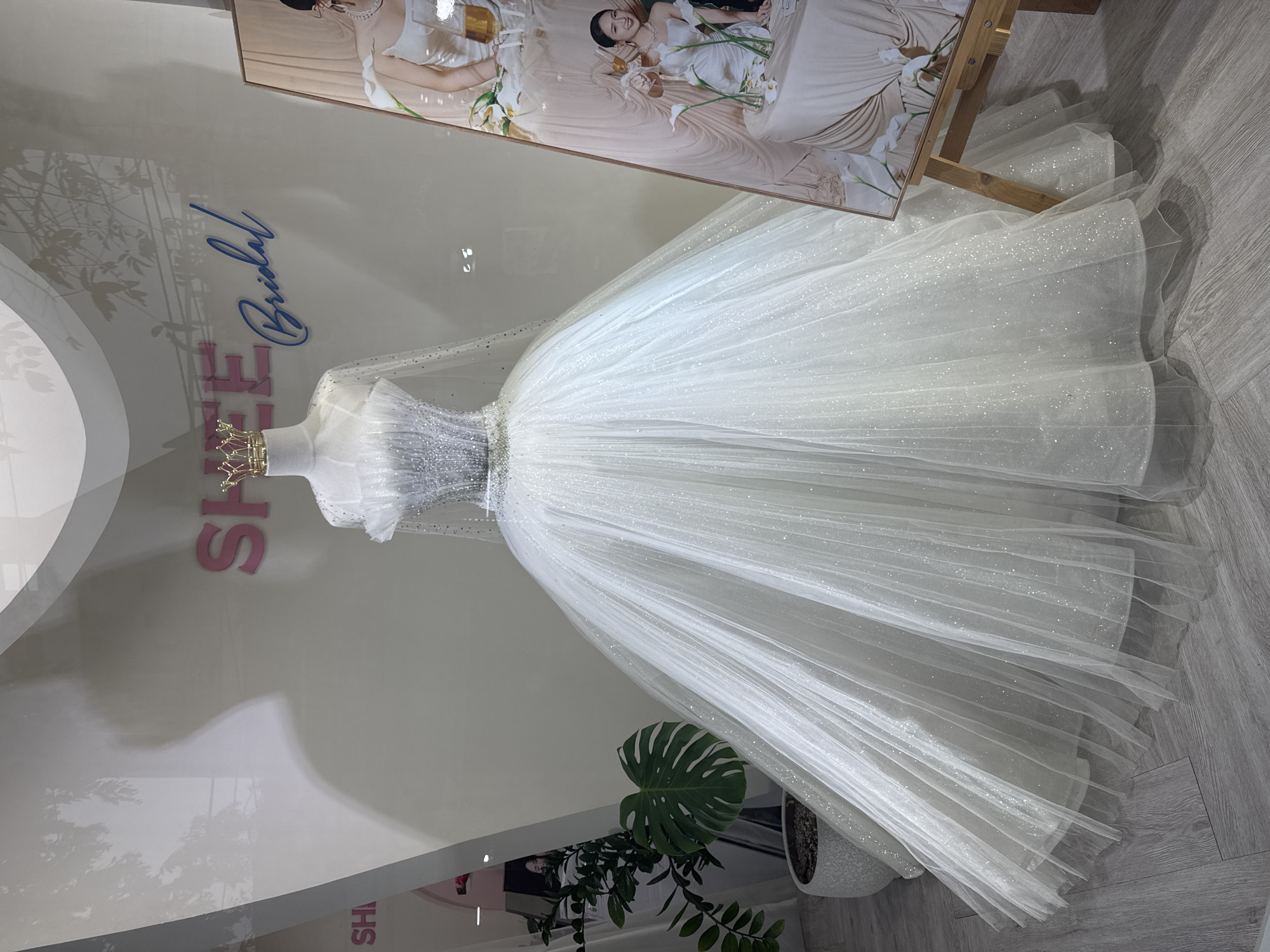 A white wedding dress on a mannequin is displayed in a shop window. The dress has a sparkly bodice and a full, tulle skirt. A small gold crown is on the mannequin's head. The shop's name, "Shee Bridal," is vertically written on the window. A framed wedding photo leans against the window behind the dress. A potted monstera plant is near the base of the dress. The scene likely takes place in Đà Nẵng, Vietnam, given the business name and general aesthetic. The environment consists of a retail setting with a bright interior.
