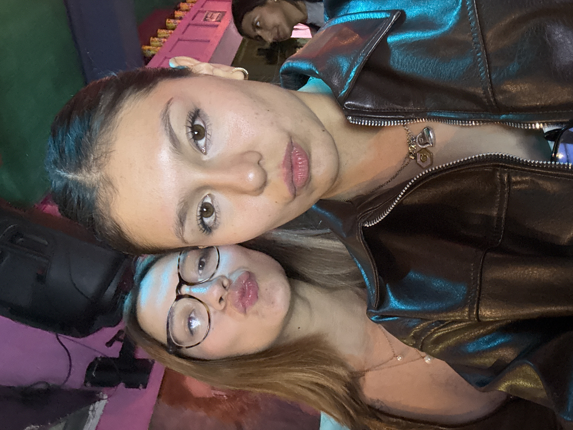 Two young women are posing closely together for a photograph, angled towards the camera. One woman has brown hair pulled back, brown eyes, and is wearing a black leather jacket. A necklace with multiple charms is visible around her neck. The other woman has longer brown hair, is wearing round tortoiseshell glasses, and is making a kissing face. A light necklace is also visible on her neck. Another person is partially visible in the background, looking down at the subjects. A pink structure is situated behind the subjects, while a speaker sits on the left. The location appears to be in San Andrés Cholula, Mexico.