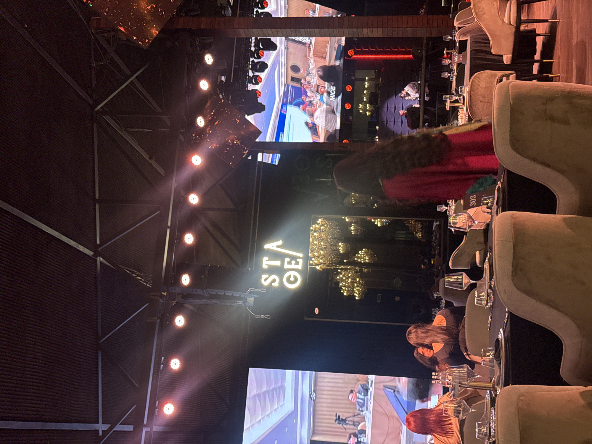 The image depicts the interior of a likely event space with high ceilings and exposed metal beams illuminated by multiple rows of lights. There is a large screen displaying a video feed, potentially a live event or conference. The word "STAGE" is illuminated vertically. The scene appears to be a gathering or banquet, with tables set for dining, complete with glassware and place settings. Several people are seated at the tables. The location is Yerevan, Armenia.