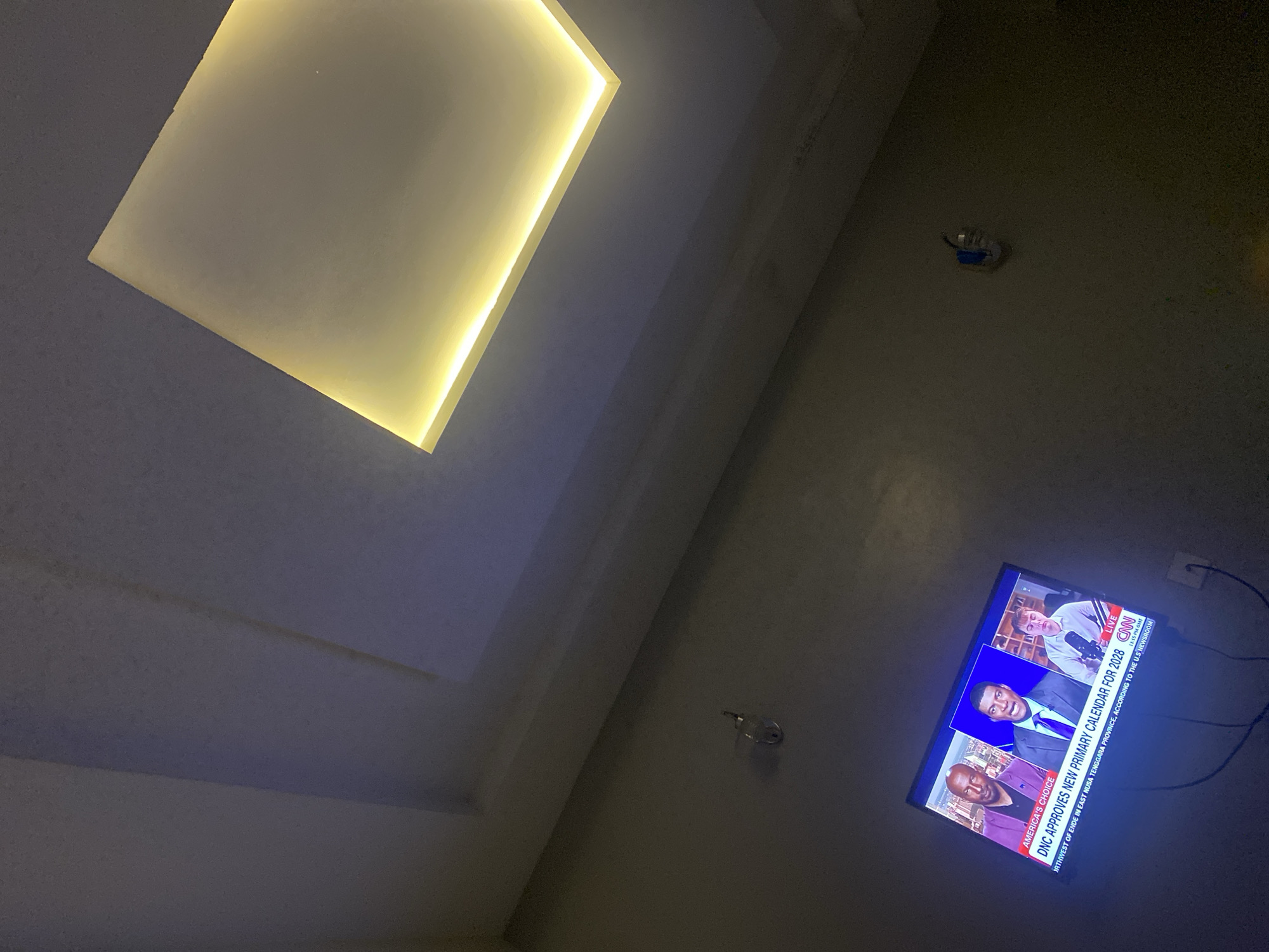 The image depicts an indoor scene, likely a living room, focused on a television screen mounted on a dark gray wall. The television displays a news broadcast from CNN, featuring three men. The man in the center, wearing a suit and tie, is speaking. Text on the screen reads "AMERICA'S CHOICE DNC APPROVES NEW PRIMARY CALENDAR FOR 2028" and "LIVE CNN". The room is dimly lit, with a prominent recessed ceiling light fixture emitting a warm yellow glow. A wall-mounted light fixture is visible on the left side of the wall. The overall impression is of a domestic setting, possibly in Barikin Jirga Summa, Nigeria, with the television serving as the primary focal point.