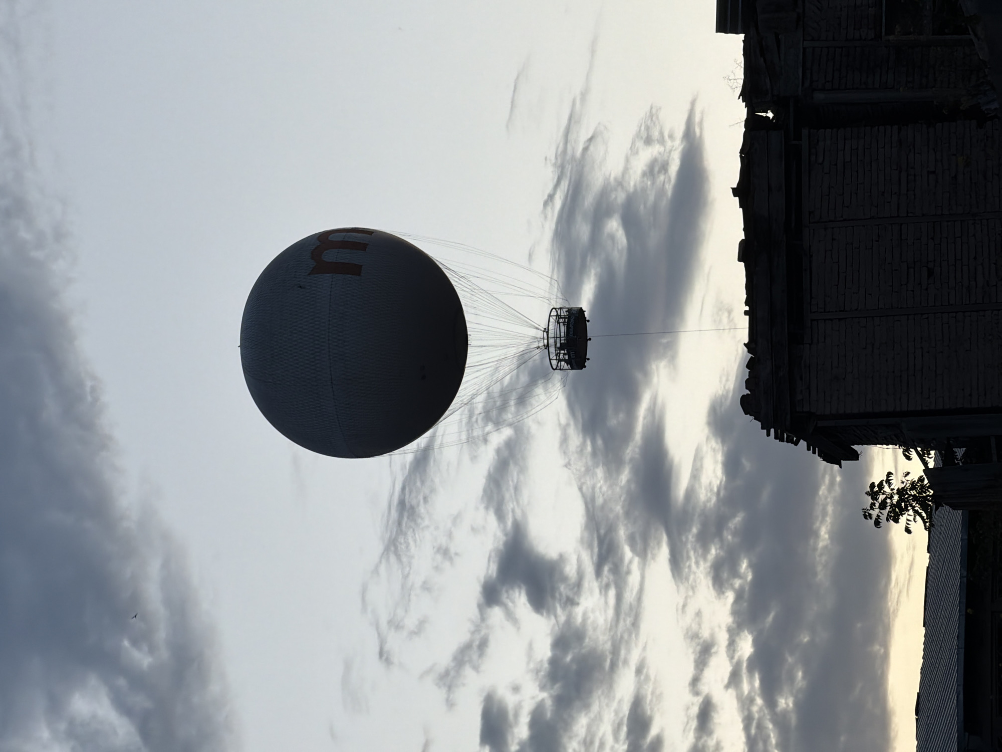 The image shows a tethered hot air balloon in Tbilisi, Georgia. The balloon is spherical, gray, and has a red letter 'M' on its side. A gondola is suspended beneath the balloon by numerous cables. The sky is overcast with gray and white clouds. A building silhouette is seen in the upper right corner.