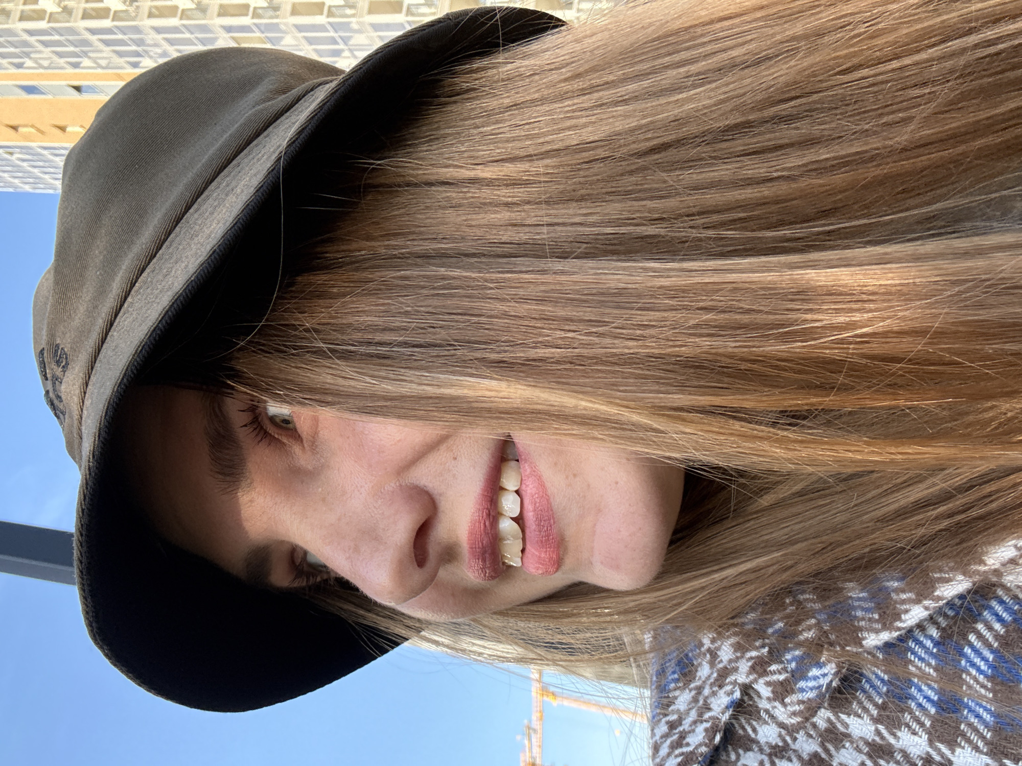 A young woman wearing a dark bucket hat and a patterned jacket is pictured in a close-up, profile shot. Her light brown hair is visible beneath the hat. She is smiling, showing her teeth, and her eyes are looking to the left. The background includes a bright blue sky, a modern building with many windows, and a construction crane, suggesting an urban setting in Kazan, Russia. The lighting indicates it is daytime.